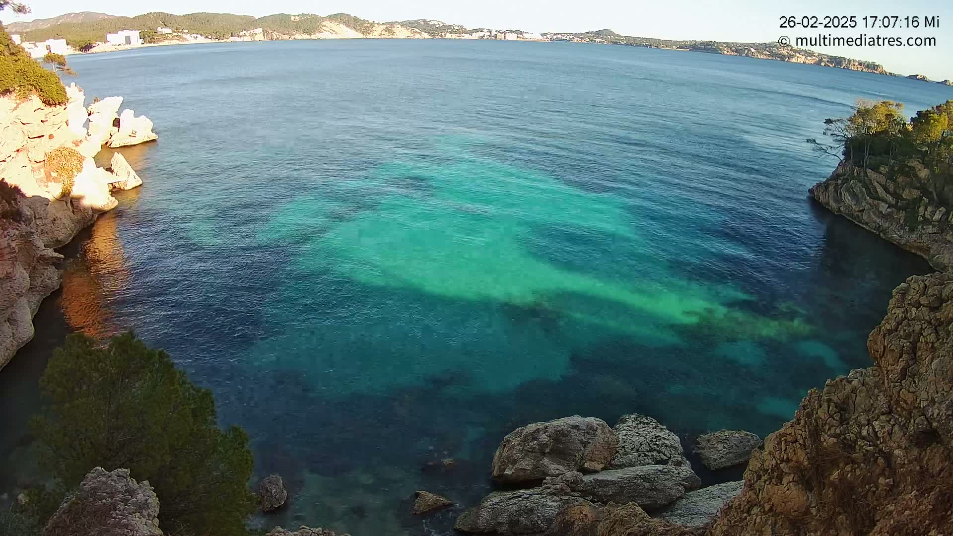 A tranquil cove with clear, teal water is nestled between rocky, vegetated cliffs under a sunny sky.