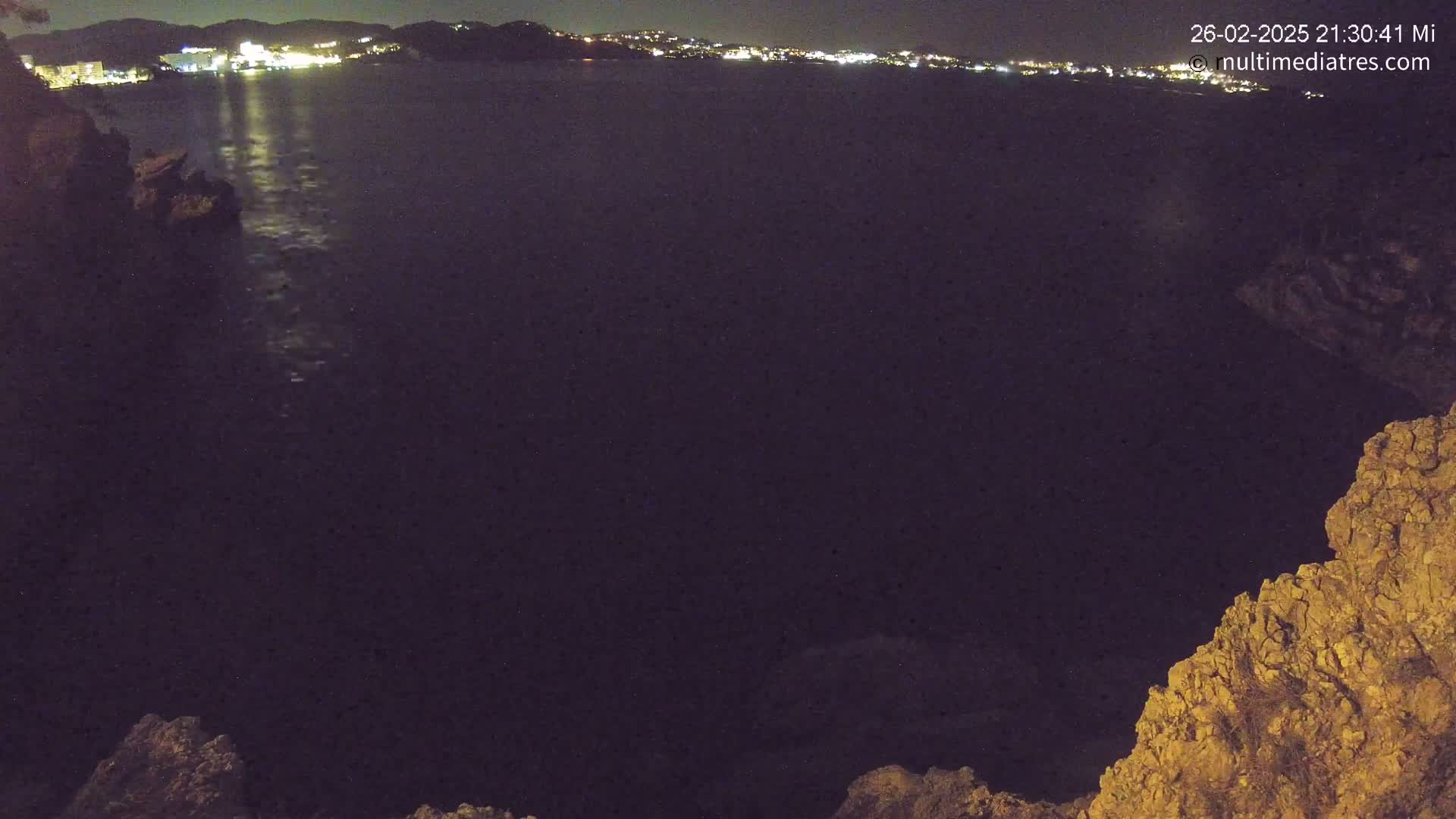 A nighttime view of a dark body of water with a distant, illuminated shoreline, partially obscured by rocky outcroppings in the foreground.