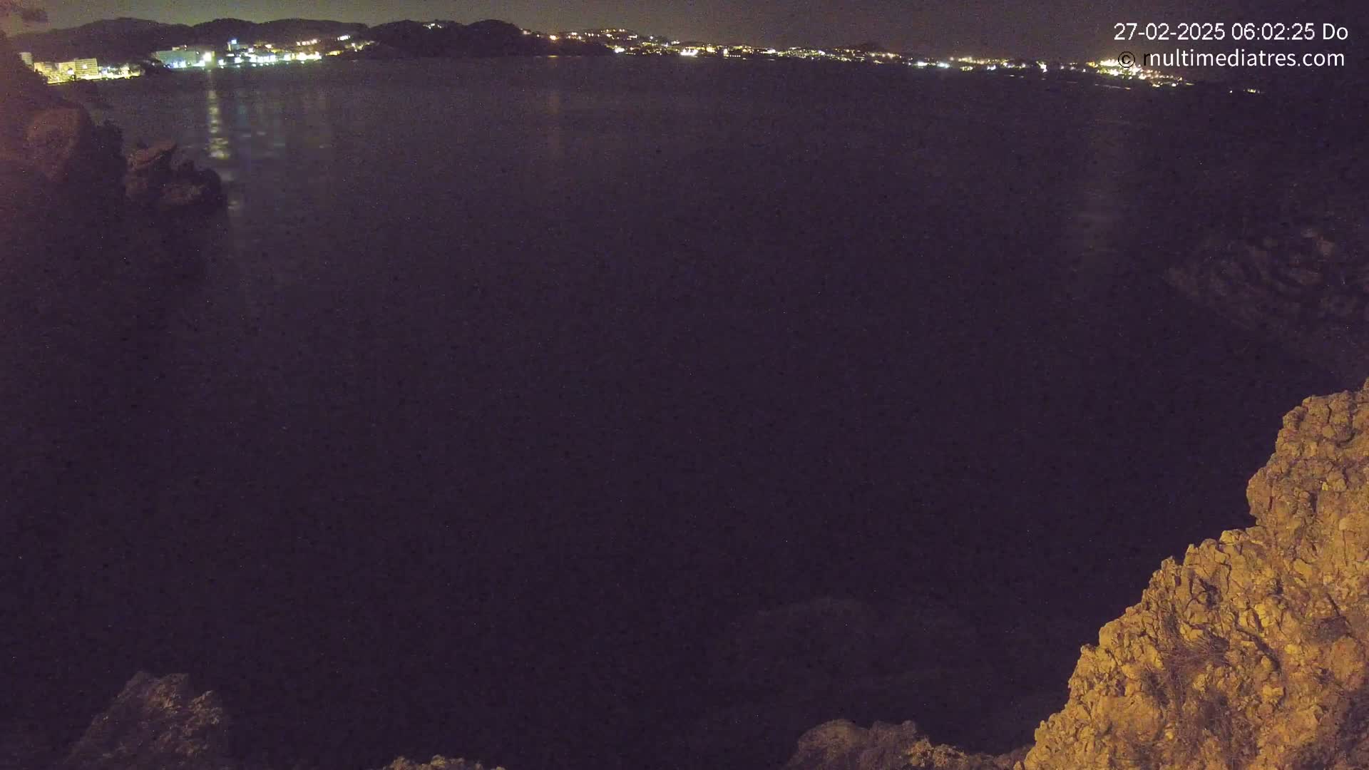 A dark, nighttime view of a calm body of water reflecting the lights of a distant town, framed by rocky terrain.