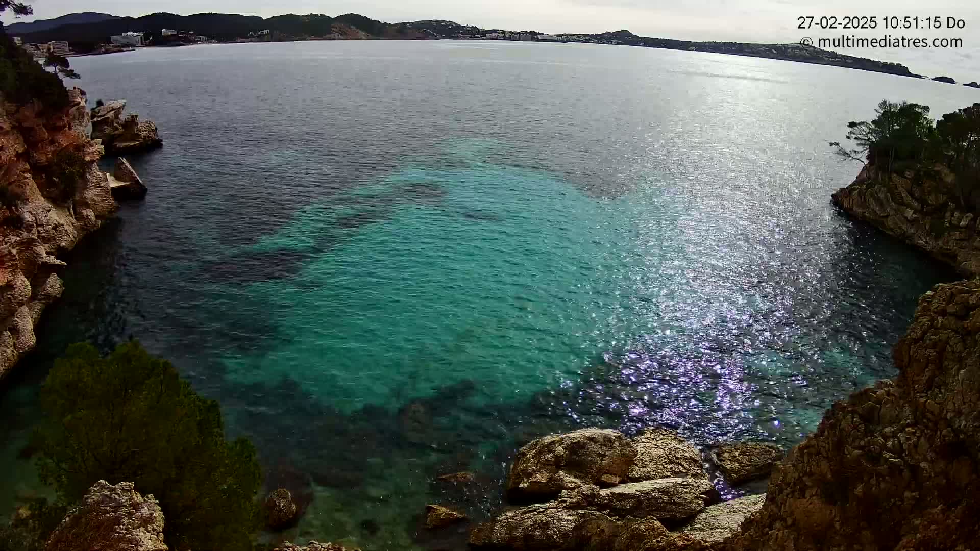 A cove of clear, turquoise water is nestled between rocky cliffs on a partly sunny day.