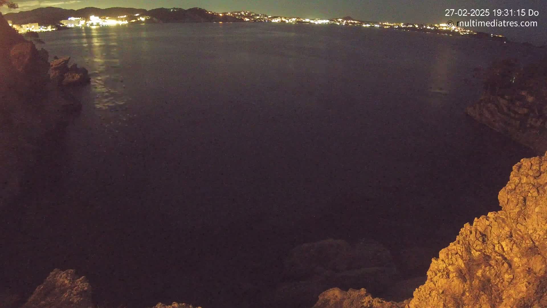 A nighttime view of a dark body of water with a distant, illuminated shoreline, seen from a rocky vantage point.