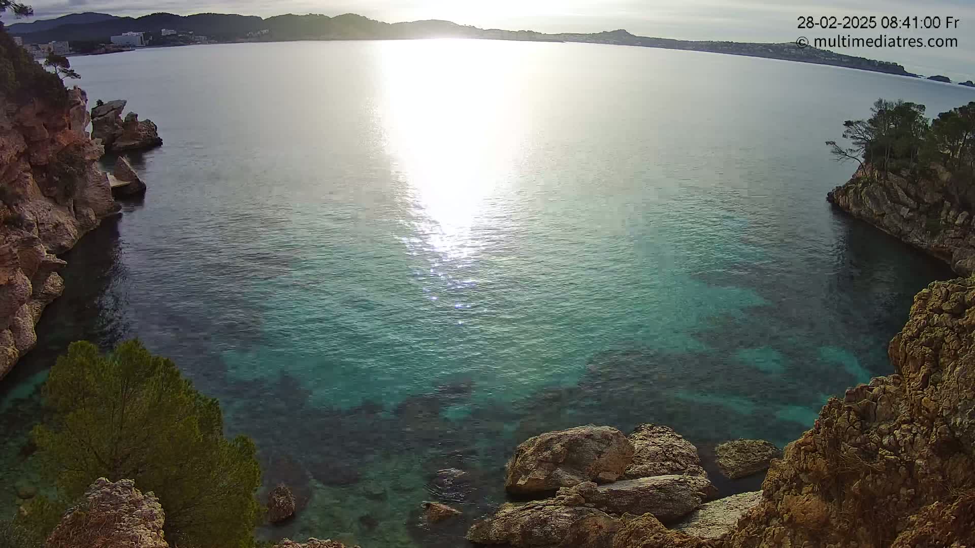 Clear, calm turquoise water is nestled within rocky, tan-colored cliffs, with a distant shoreline visible under a bright, sunny sky.