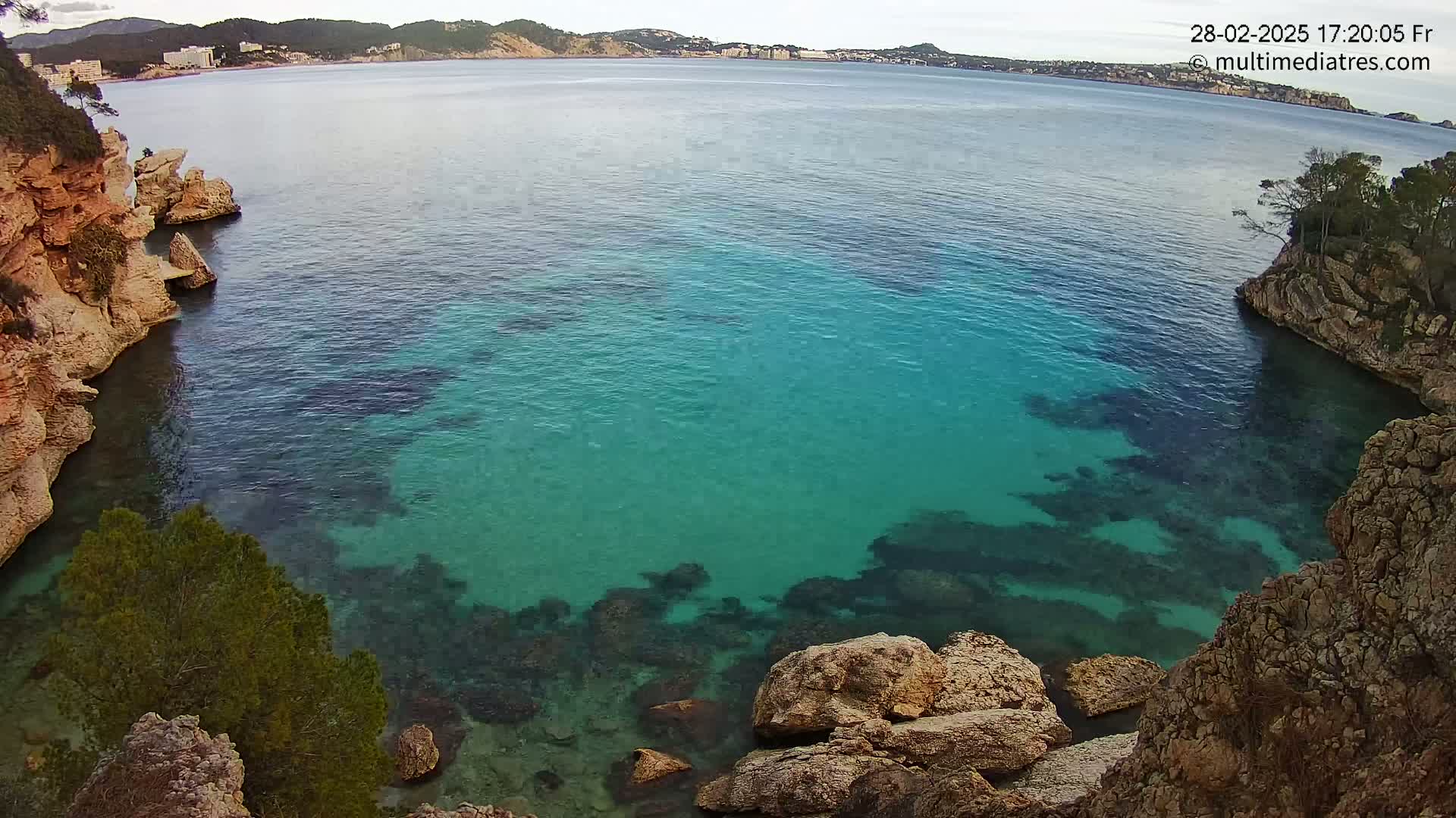 A cove with clear, teal water is nestled between rocky, tan-colored cliffs under a mostly cloudy sky.