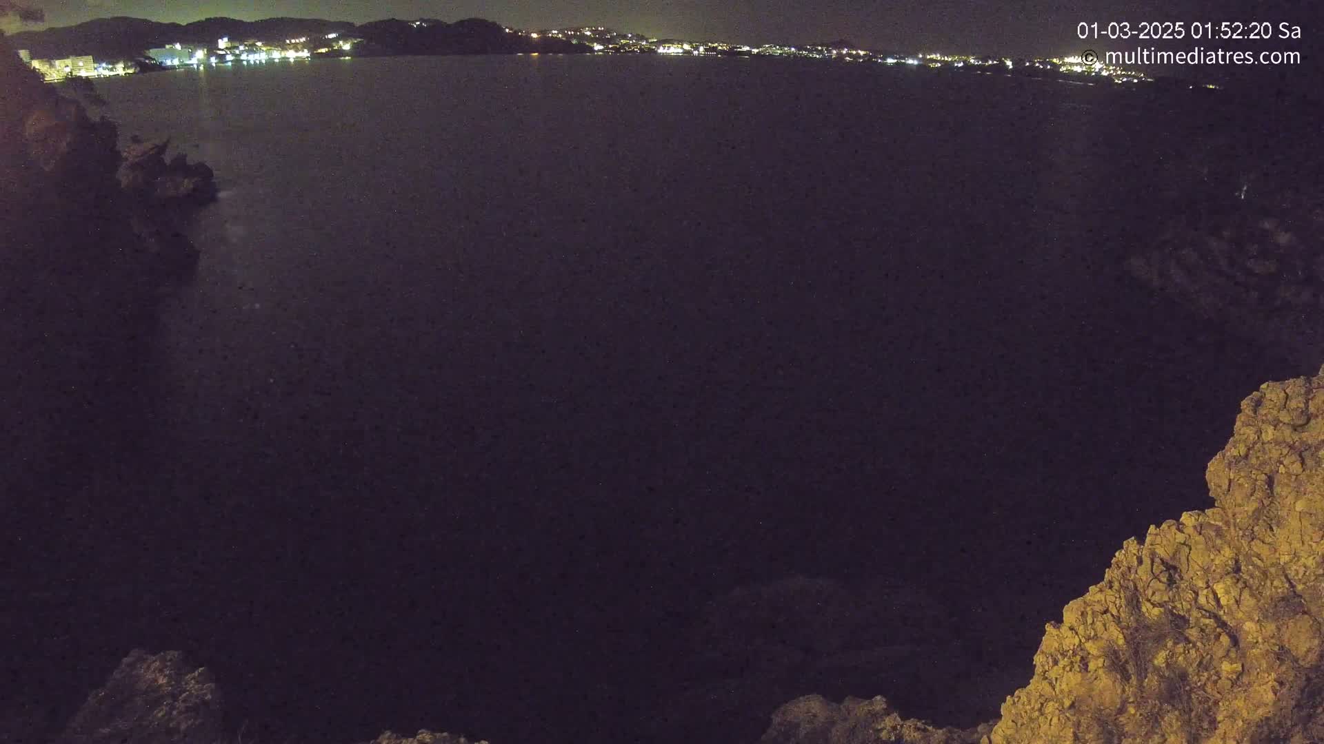 A dark, nighttime view of a calm body of water, with a distant cityscape of lights visible on the horizon, framed by rocky terrain.
