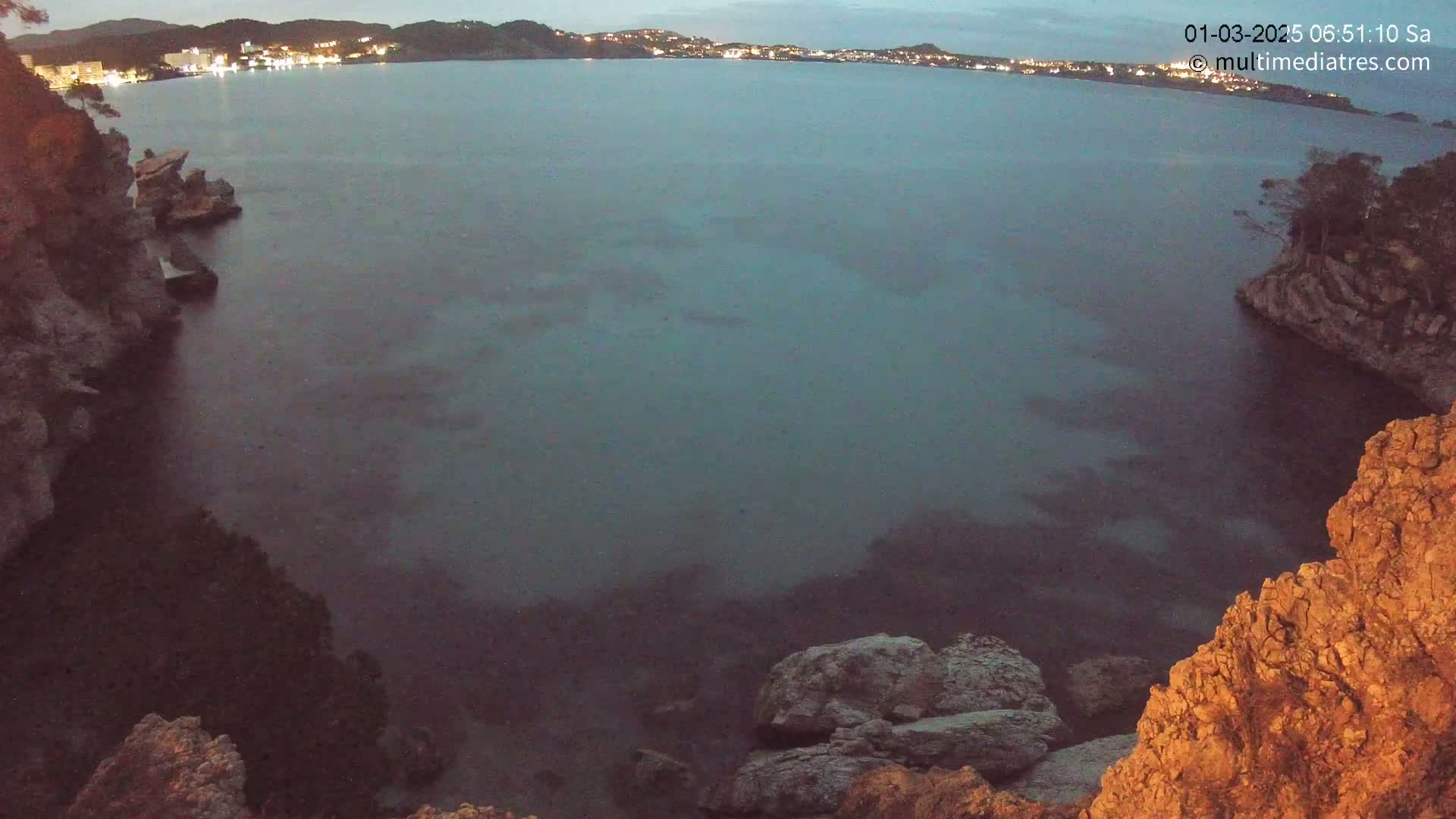 A dark, calm body of water is nestled between rocky cliffs at twilight, with a distant town faintly illuminated on the horizon.