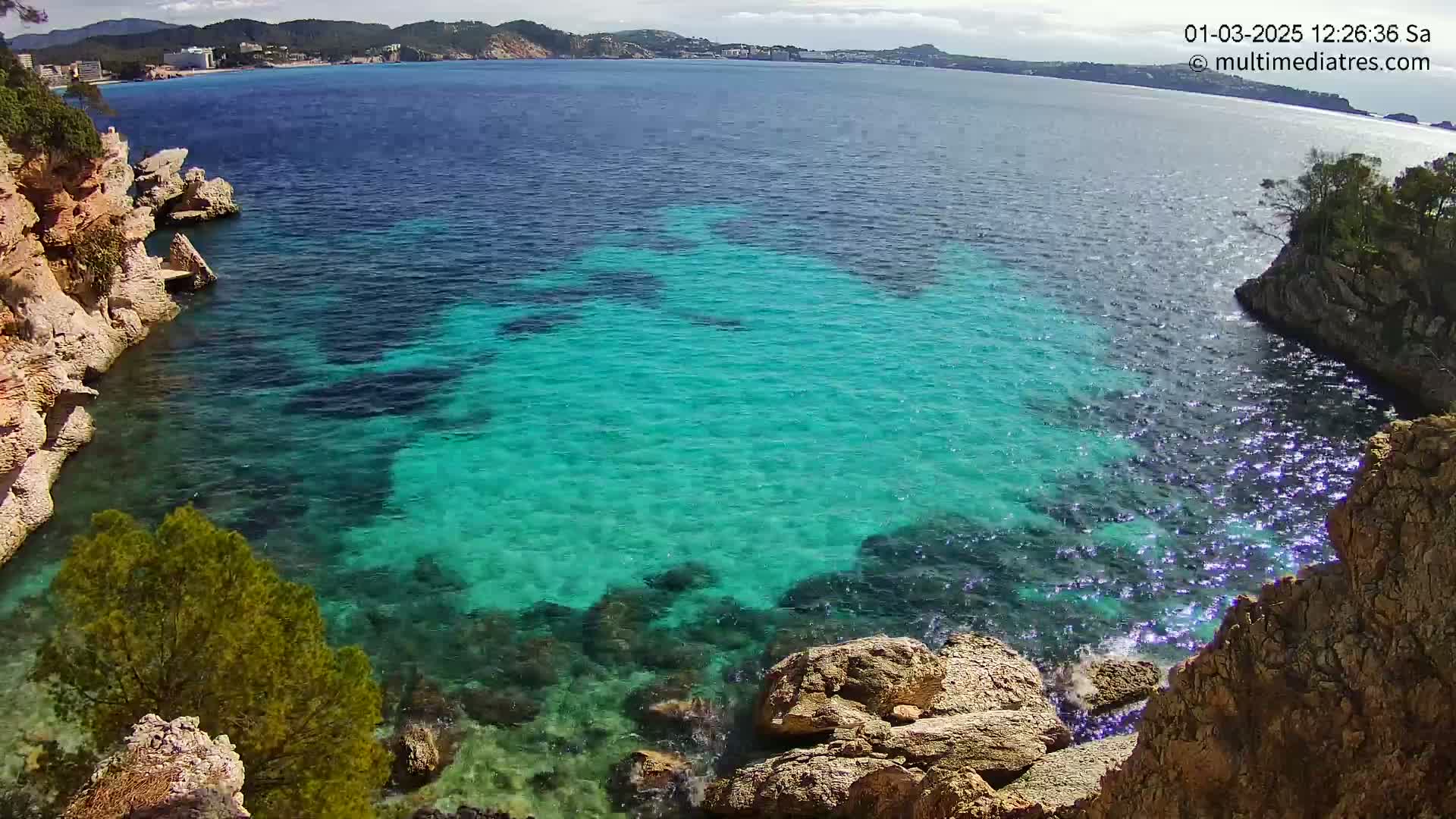 Brightly lit, clear turquoise water fills a cove bordered by light-brown rocky cliffs and vegetation under a partly sunny sky.