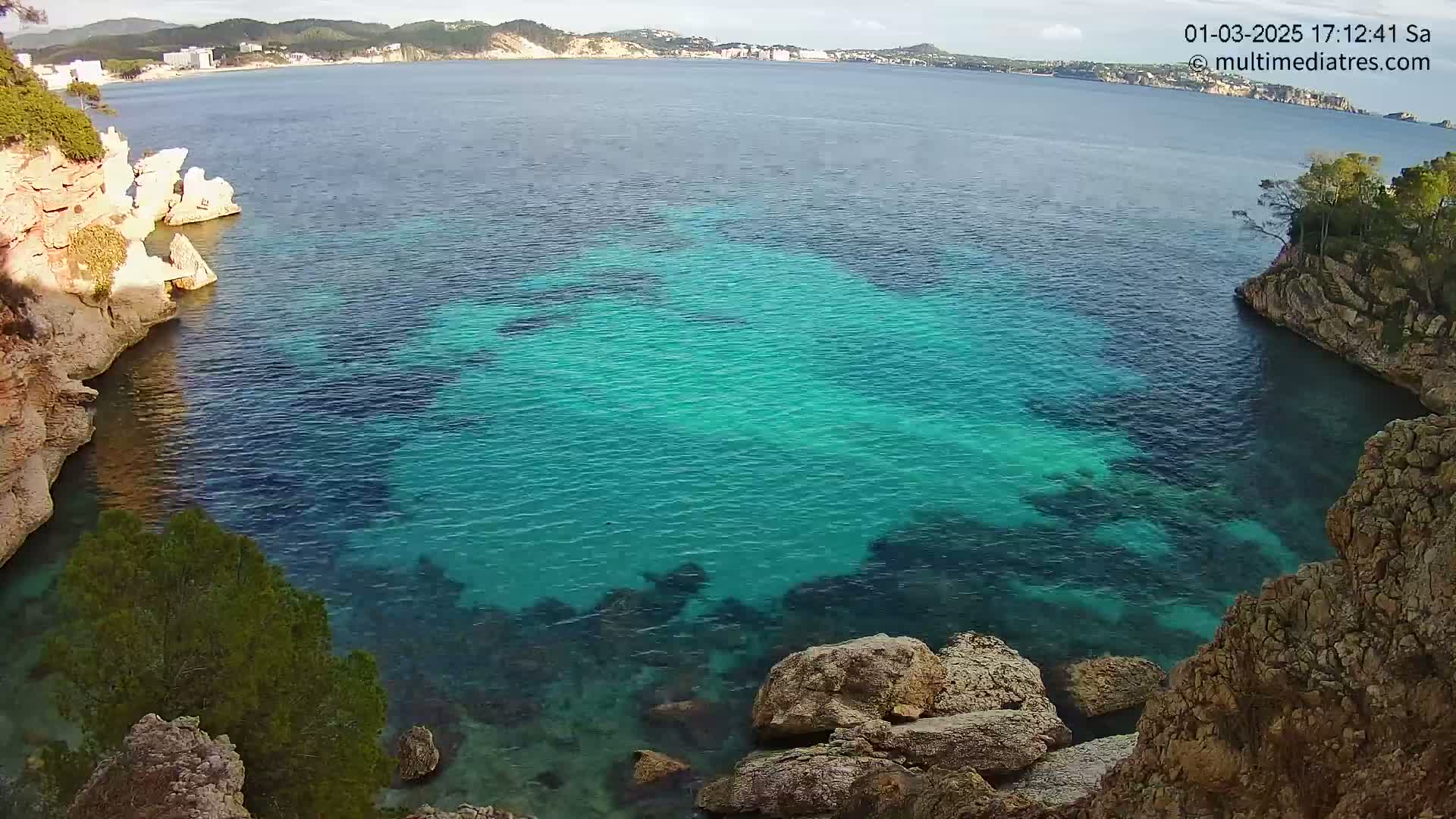 A cove of clear, teal water is nestled between rocky, vegetated cliffs under a mostly sunny sky.