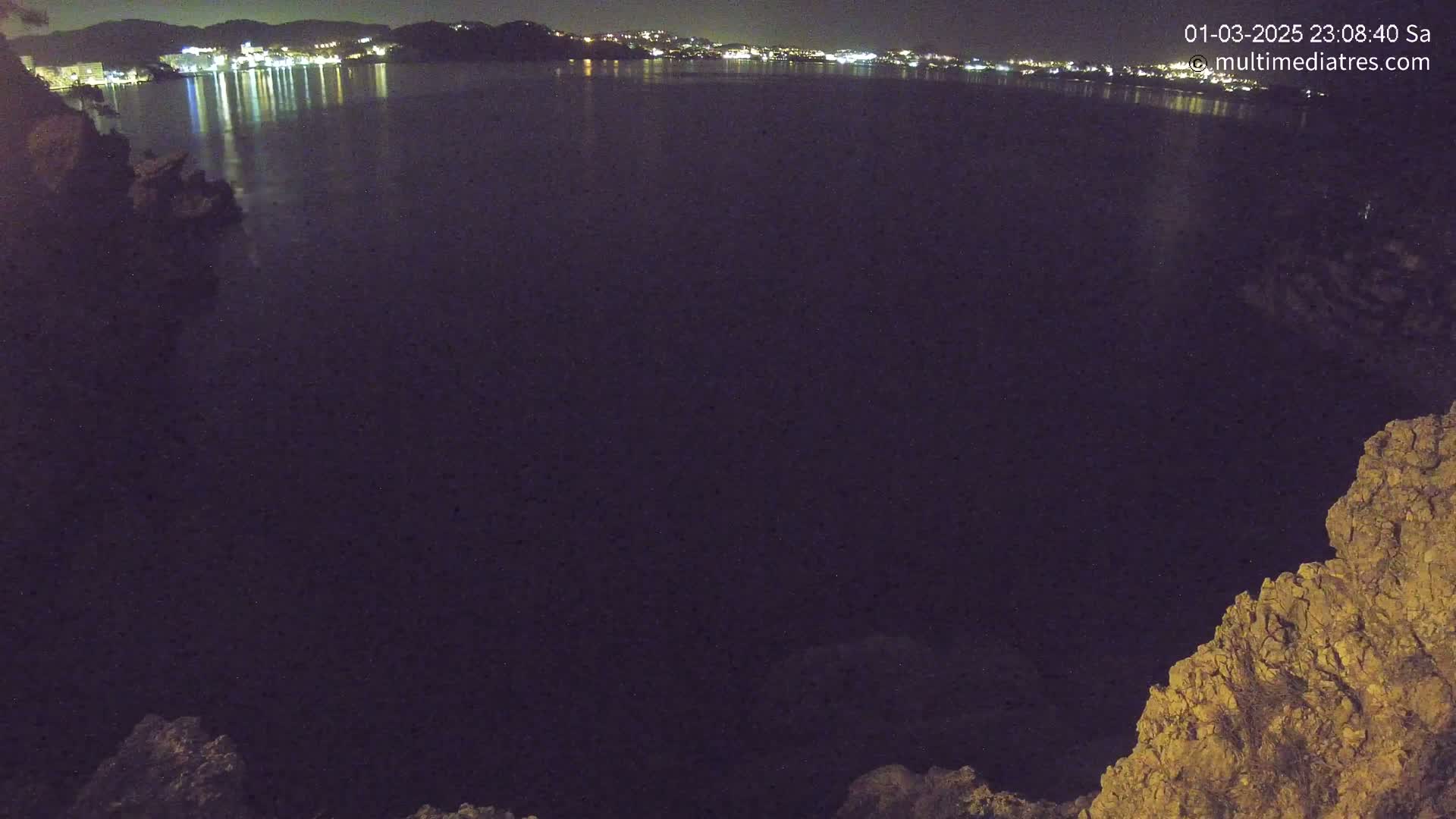 A nighttime view of a dark body of water reflecting the lights of a town across the shore, partially framed by dark rocky outcrops.