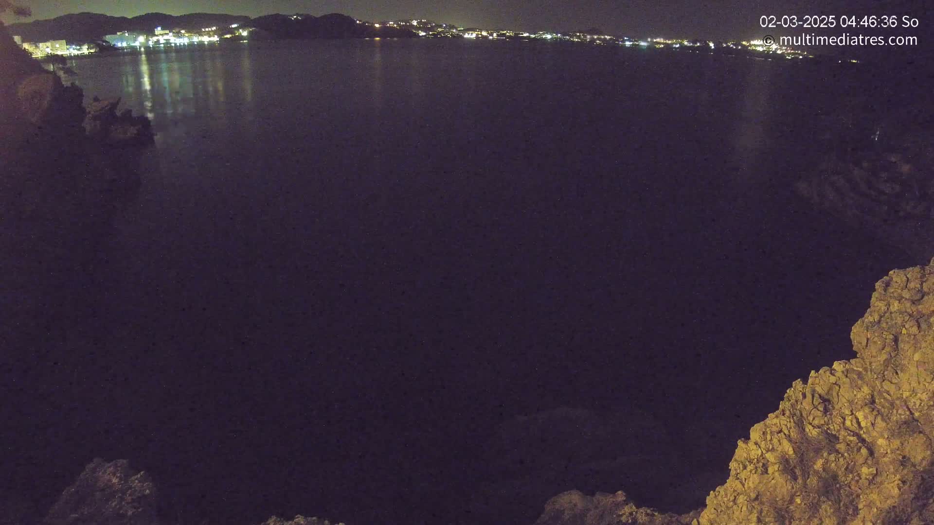 A nighttime view of a dark body of water reflecting the lights of a distant town, framed by rocky terrain.