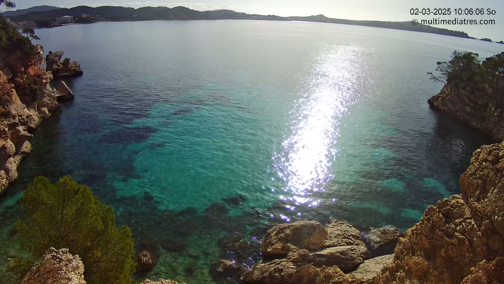 Bright sunlight glints off clear, turquoise water nestled in a rocky cove, with hills visible in the distance under a mostly sunny sky.