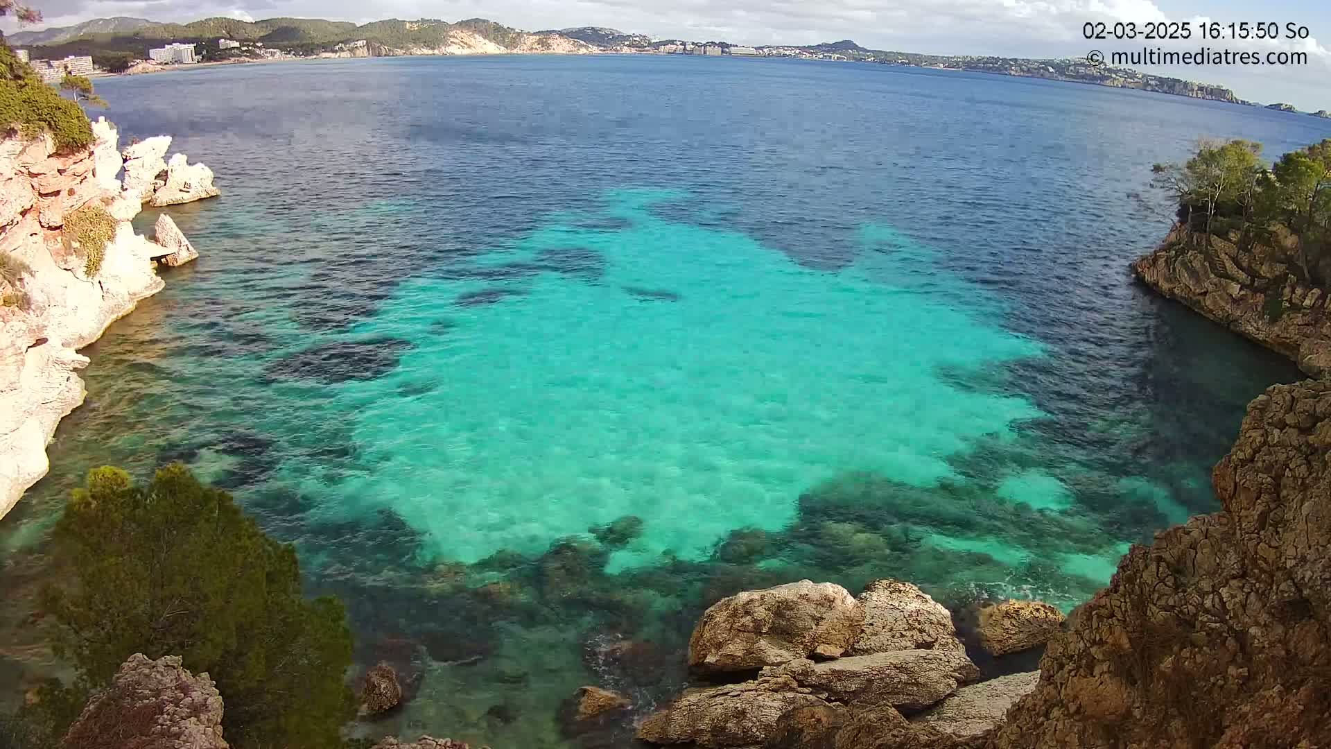 A cove of brilliantly turquoise water is nestled between light-colored rocky cliffs on a partly cloudy day.