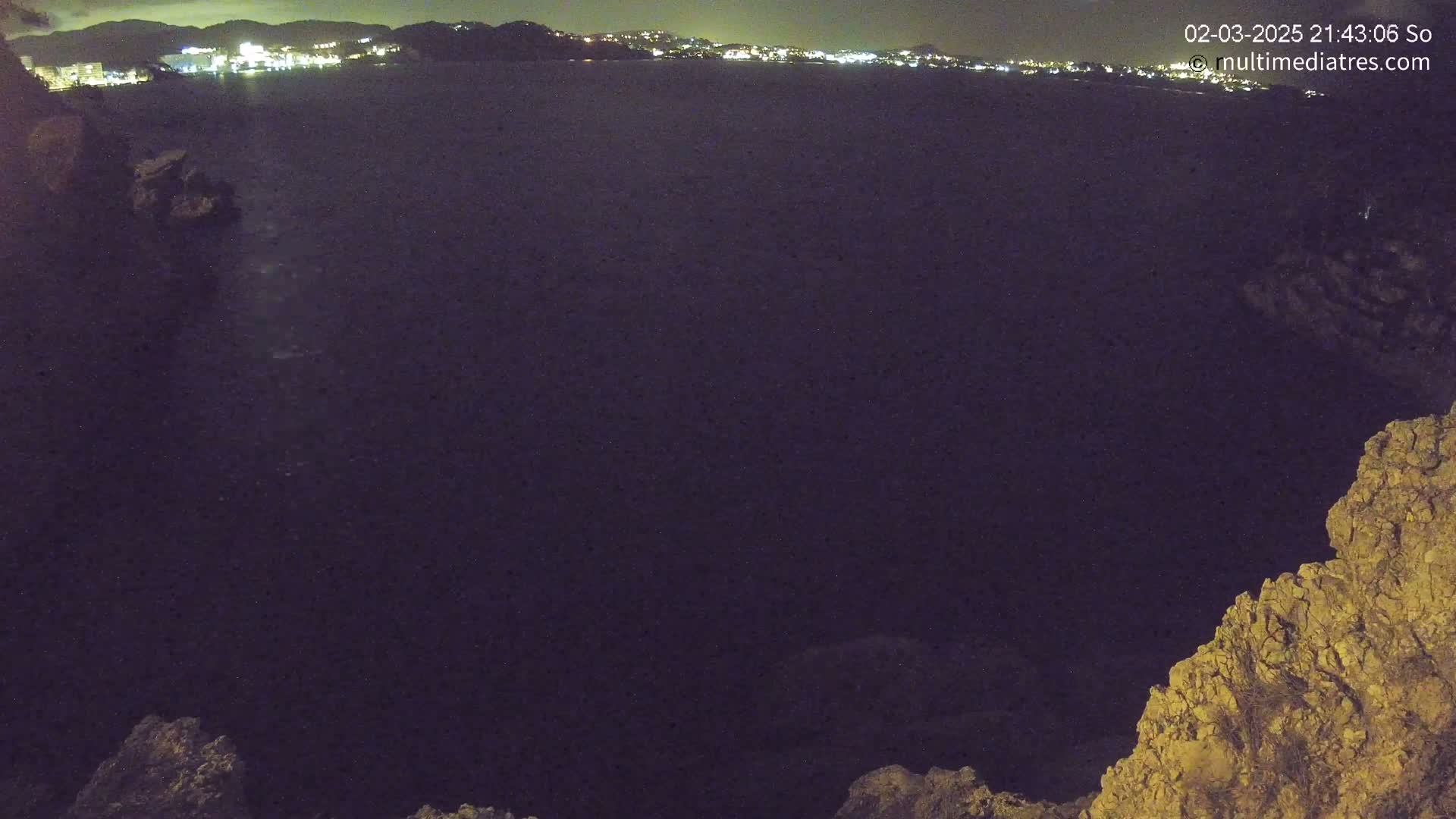 A nighttime, long-range view shows a dark body of water with a distant, illuminated town on the horizon, partially framed by rocky terrain in the foreground.