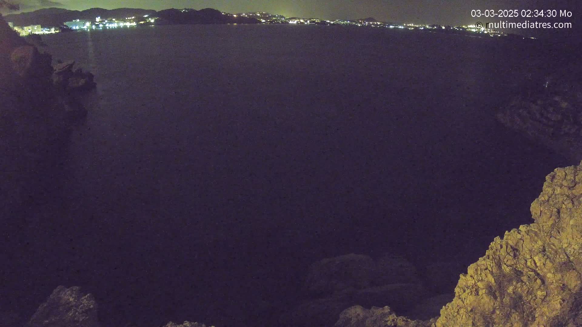 A nighttime view of a dark body of water with a distant town's lights visible across the water, partially framed by rocky terrain in the foreground.