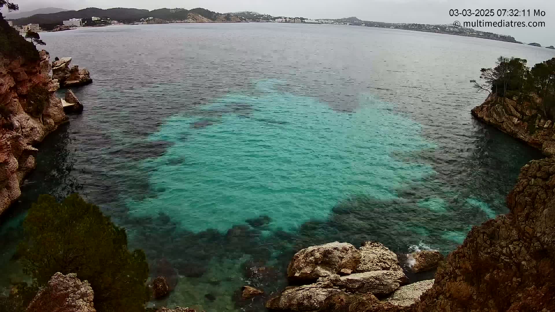 A cove of teal-colored water is surrounded by rocky, tan cliffs and greenery under an overcast sky.