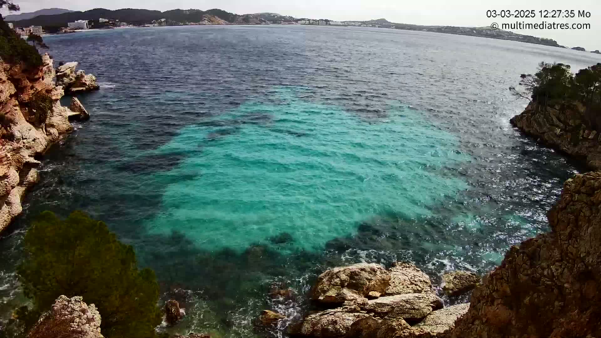 Turquoise water laps against rocky, light brown cliffs under a mostly clear sky.