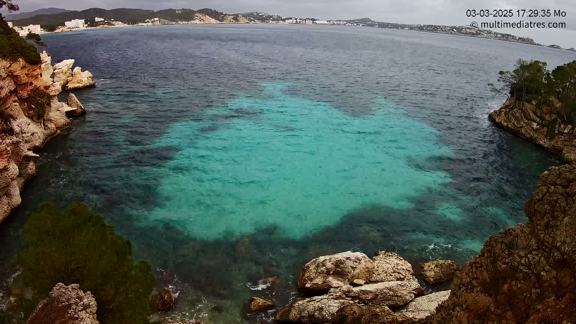 A cove of teal-colored water is nestled between rocky cliffs on an overcast day, with a distant shoreline visible across the bay.