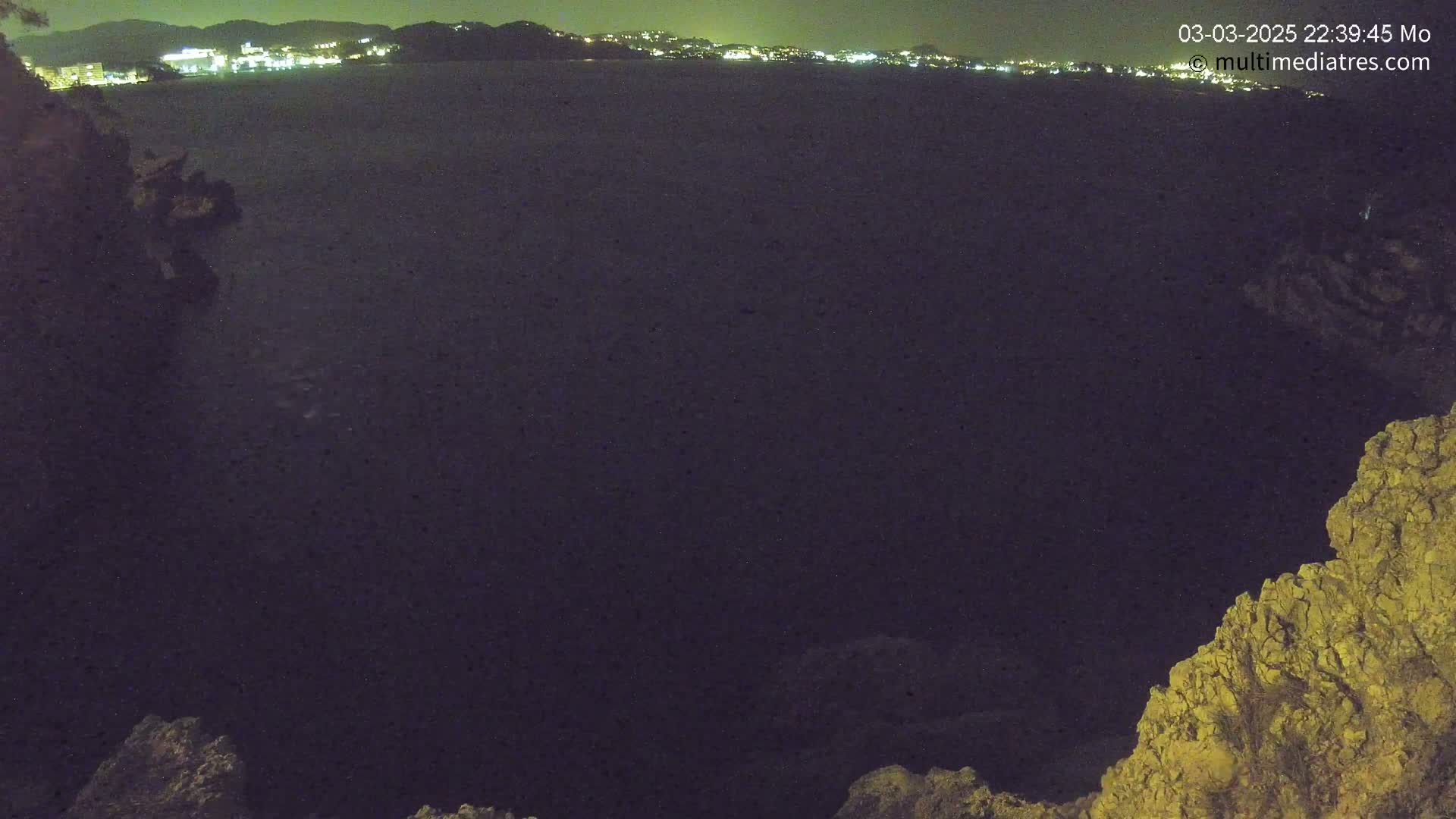 A dark, nighttime view of a calm body of water with a distant, illuminated shoreline, seen from a rocky overlook.