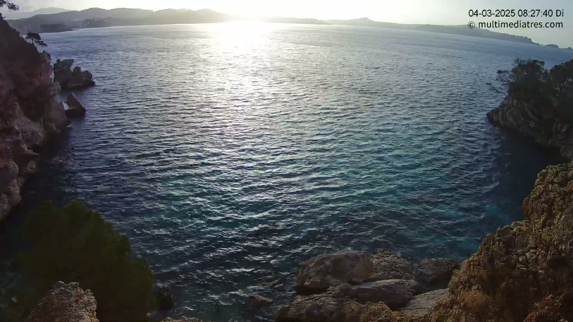 A tranquil cove of dark blue water is surrounded by rocky cliffs and vegetation, bathed in the soft light of a setting sun.