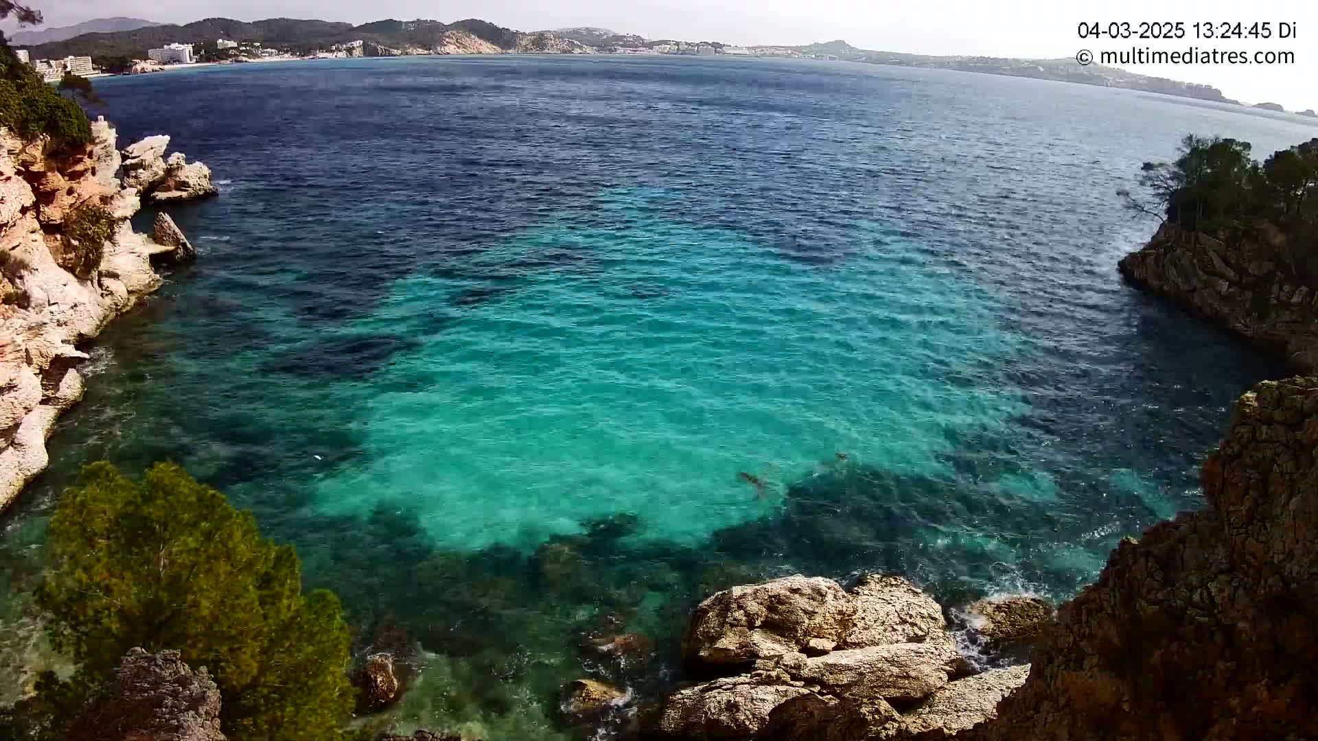 Clear, turquoise water fills a cove between rocky, vegetated cliffs on a sunny day, with a distant coastline visible in the background.