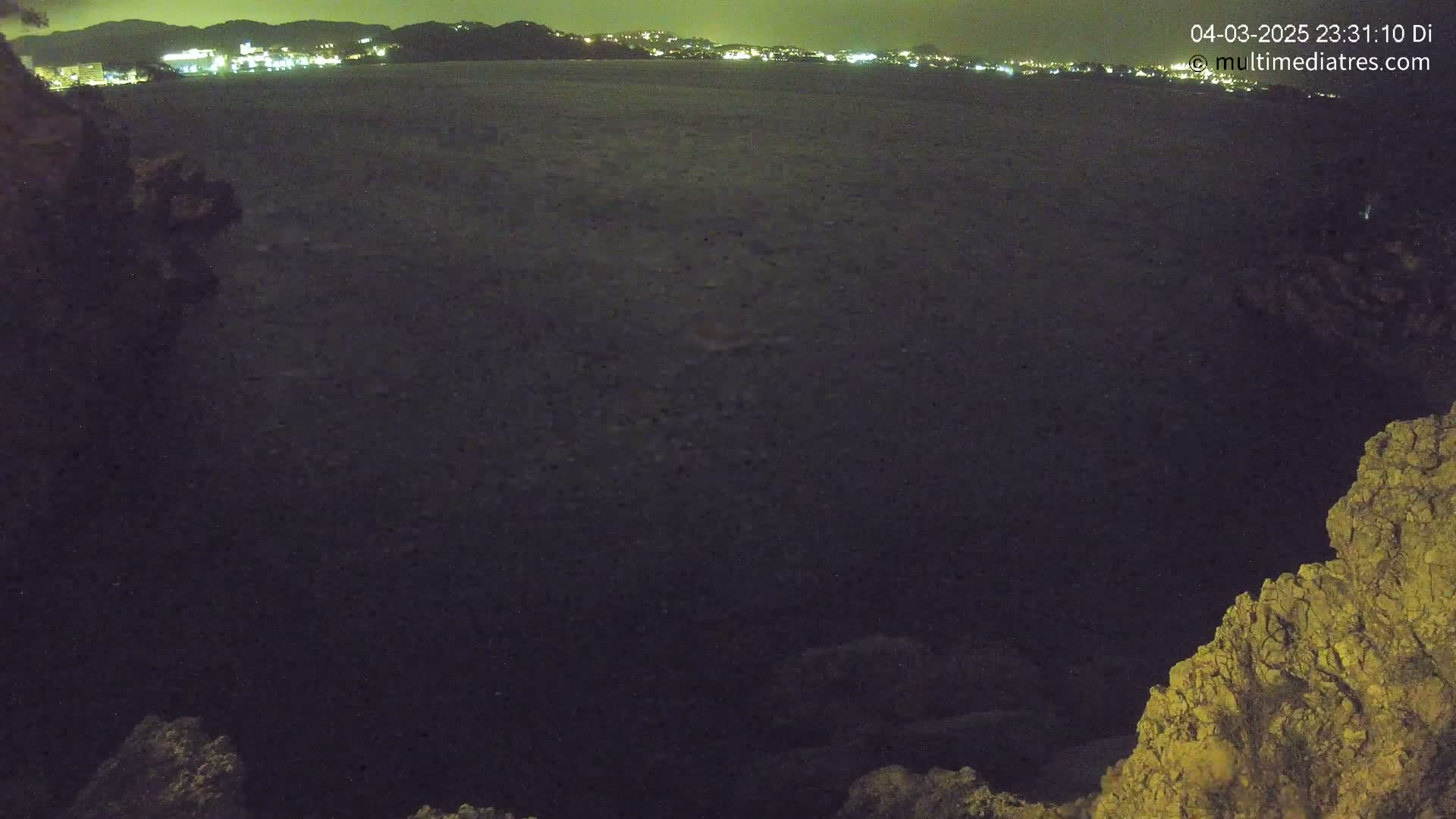 A dark body of water at night is seen from a high vantage point, with a distant, faintly lit shoreline visible in the background.