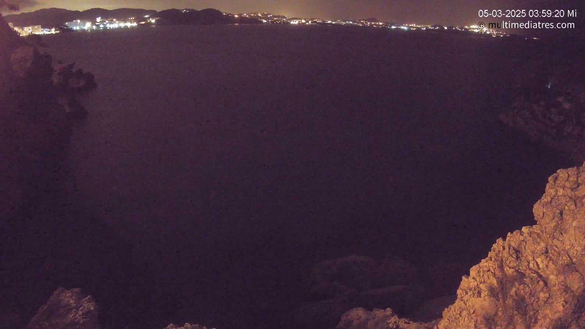 A nighttime view of dark water with a distant shoreline of city lights visible, partially framed by dark rocky outcrops.