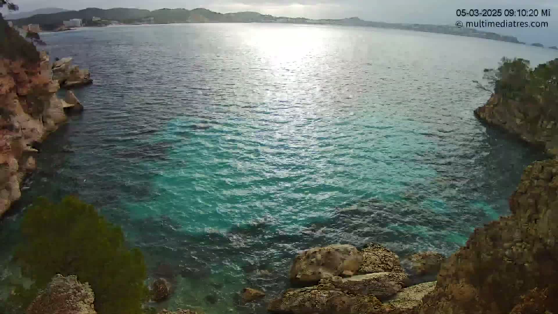 A partly sunny day shows turquoise water nestled between rocky cliffs, with a distant shoreline visible in the background.