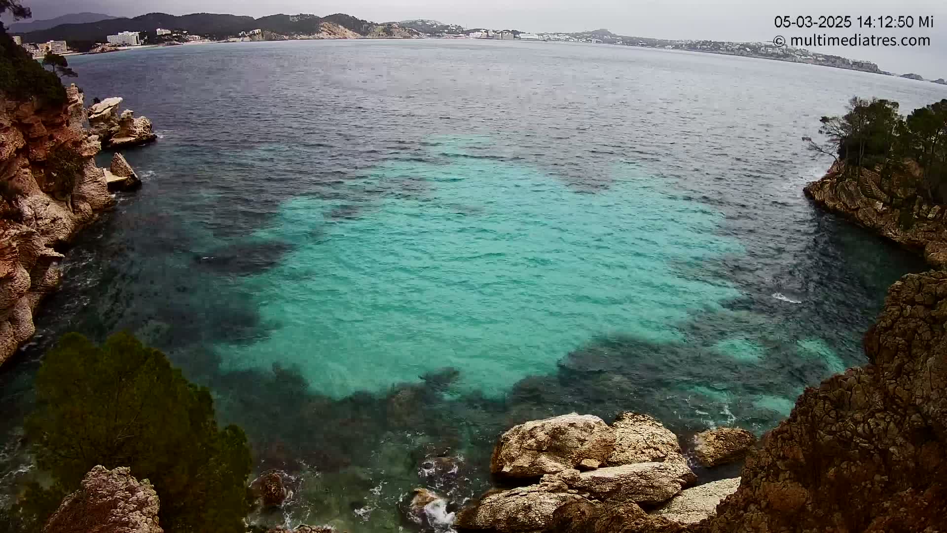 A cove of clear, teal water is nestled between rocky, tan cliffs under an overcast sky.