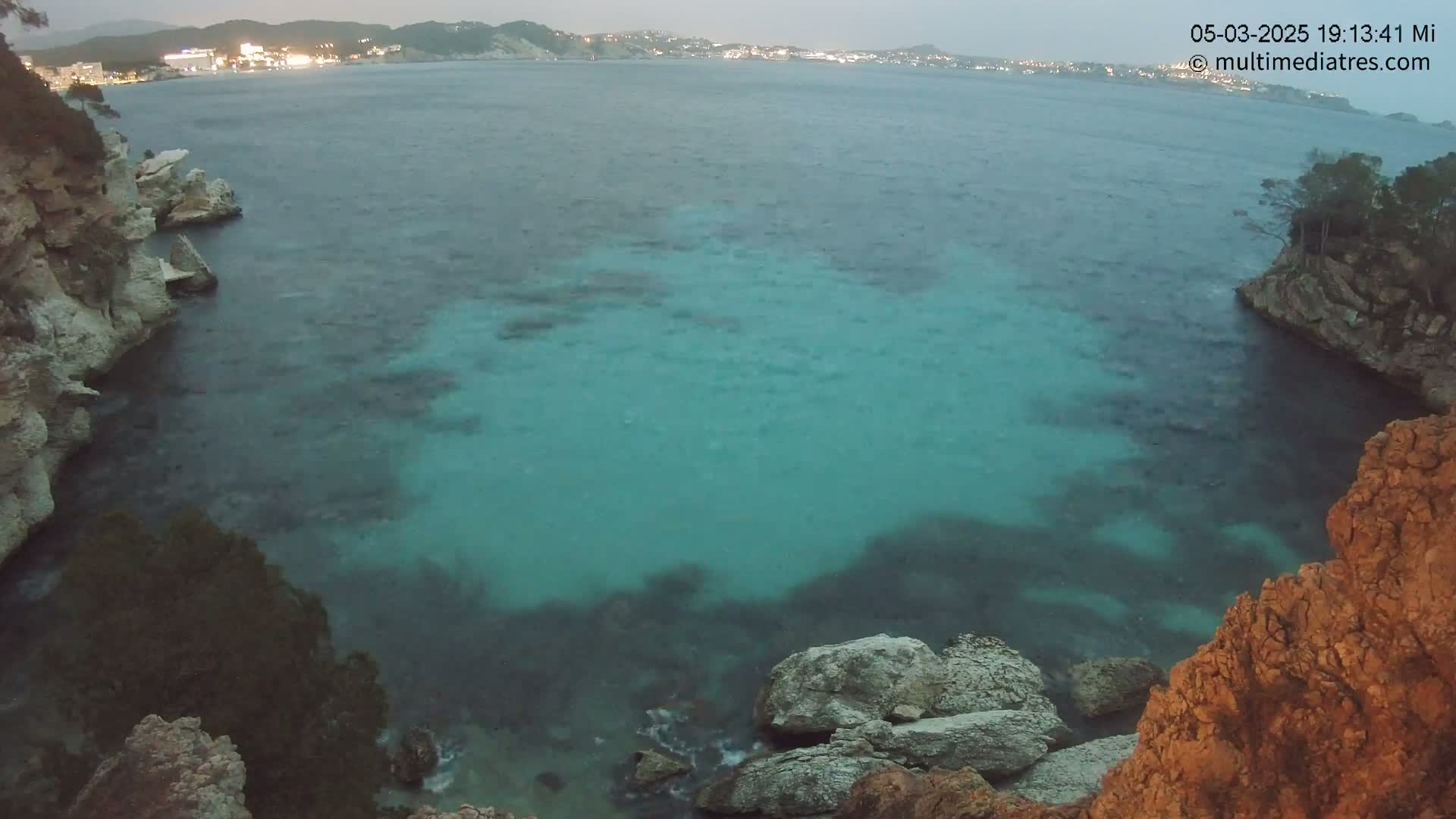 A tranquil cove with teal water is nestled between rocky cliffs at dusk, overlooking a city on the horizon.