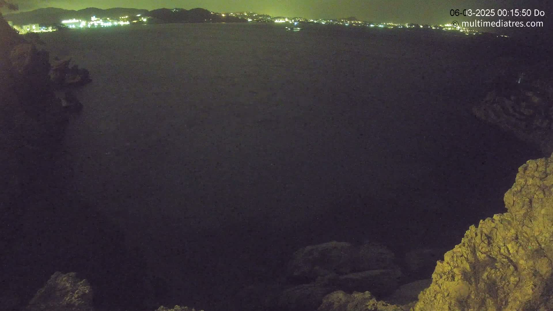 A dark, nighttime view of a body of water with a distant, lit shoreline, seen from a rocky vantage point.