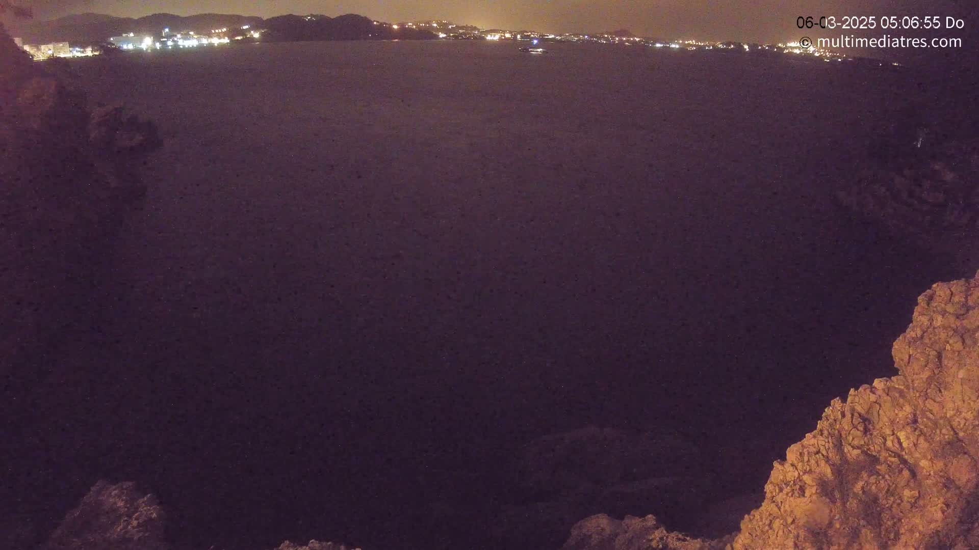 A nighttime, dark-purple-hued view of a calm body of water with a distant city skyline illuminated against a dark sky, framed by rocky outcrops in the foreground.