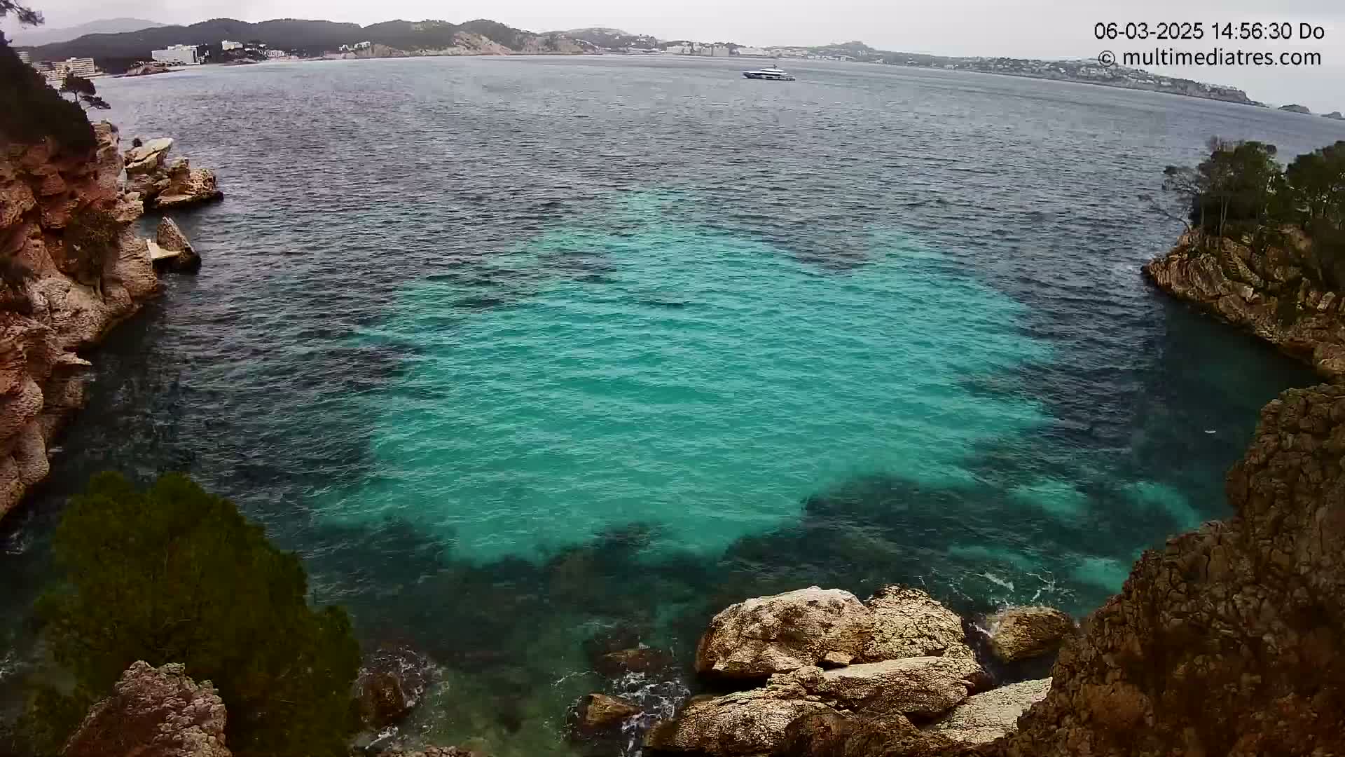 A cove of teal-colored water is nestled between rocky, tan cliffs on an overcast day, with a distant coastline visible.