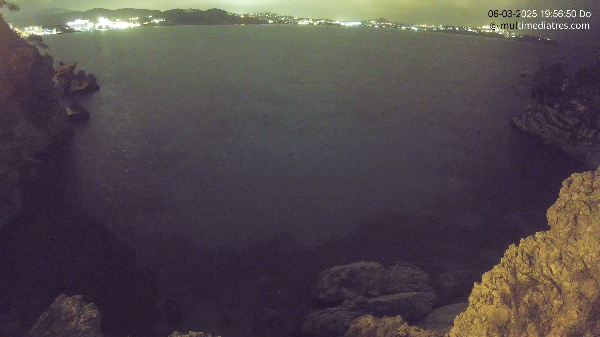 A nighttime, dark view of a calm body of water with rocky outcrops in the foreground and a distant, lit shoreline.