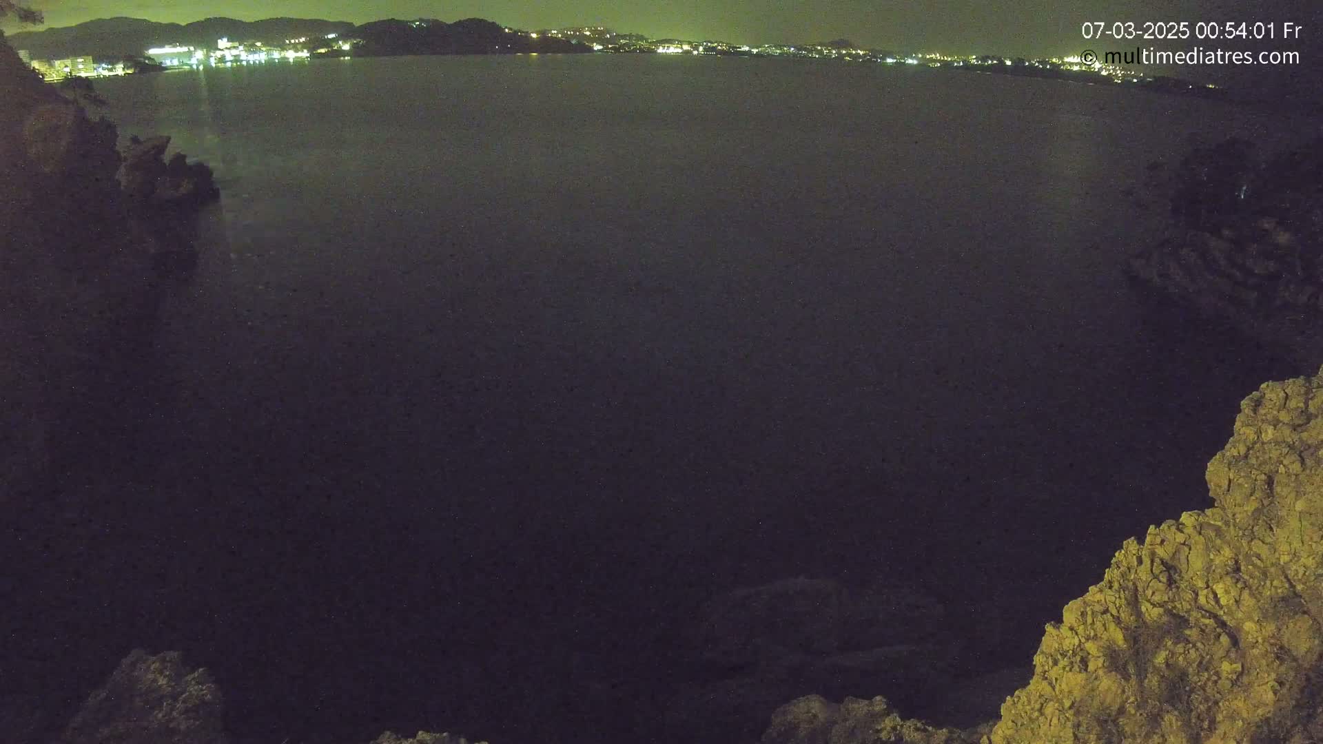 A nighttime, dark view of a calm body of water with a distant, illuminated city skyline visible in the background, partially framed by dark, rocky land.