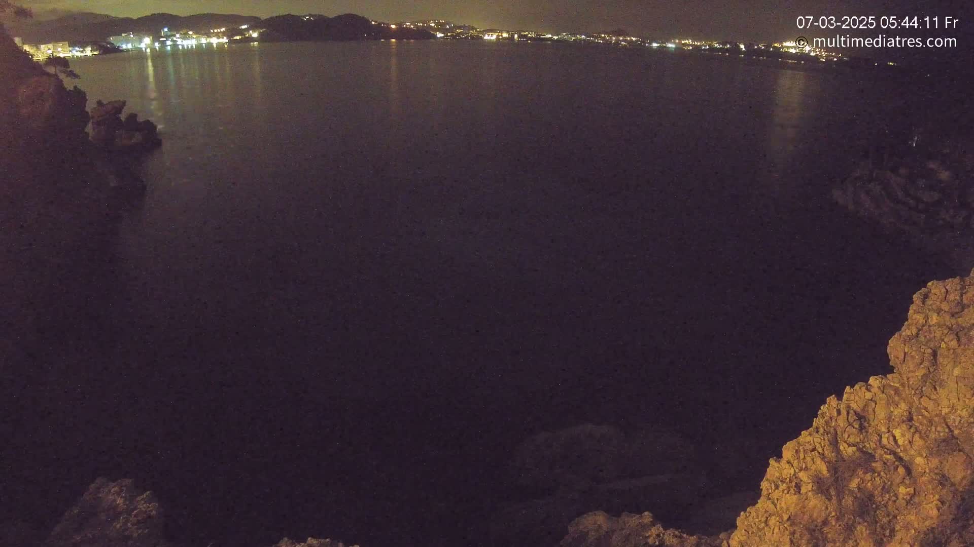 A dark body of water at night is seen from a rocky overlook, with a distant city skyline faintly illuminated.