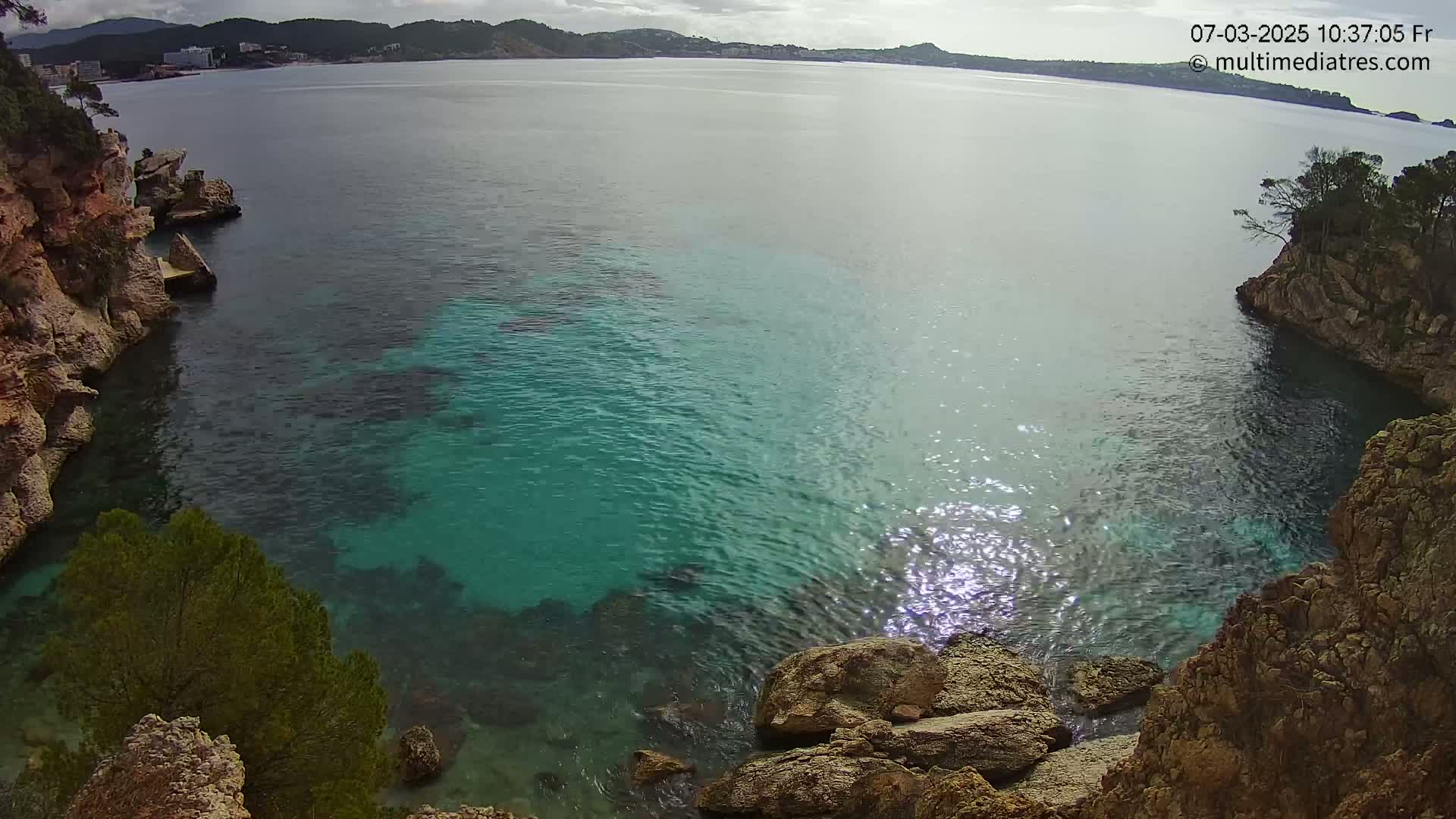 A cove with clear, teal water is nestled between rocky cliffs, with a distant coastline visible under a partly cloudy sky.