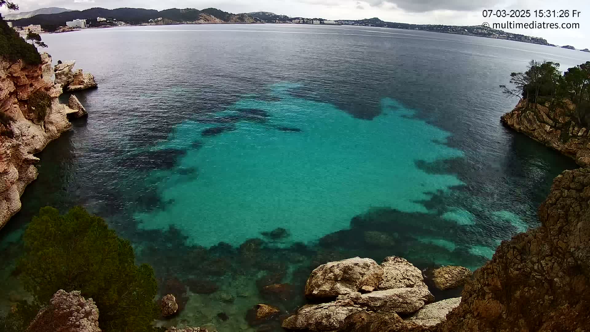 A cove with clear, teal water is surrounded by rocky cliffs and overlooks a calm sea under a cloudy sky.