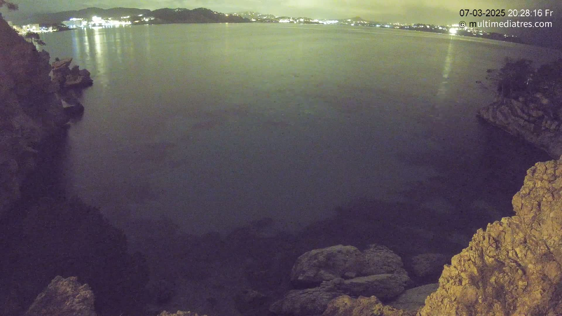 A nighttime view of a dark, calm body of water nestled between rocky shorelines, with a faintly lit town visible in the distance.