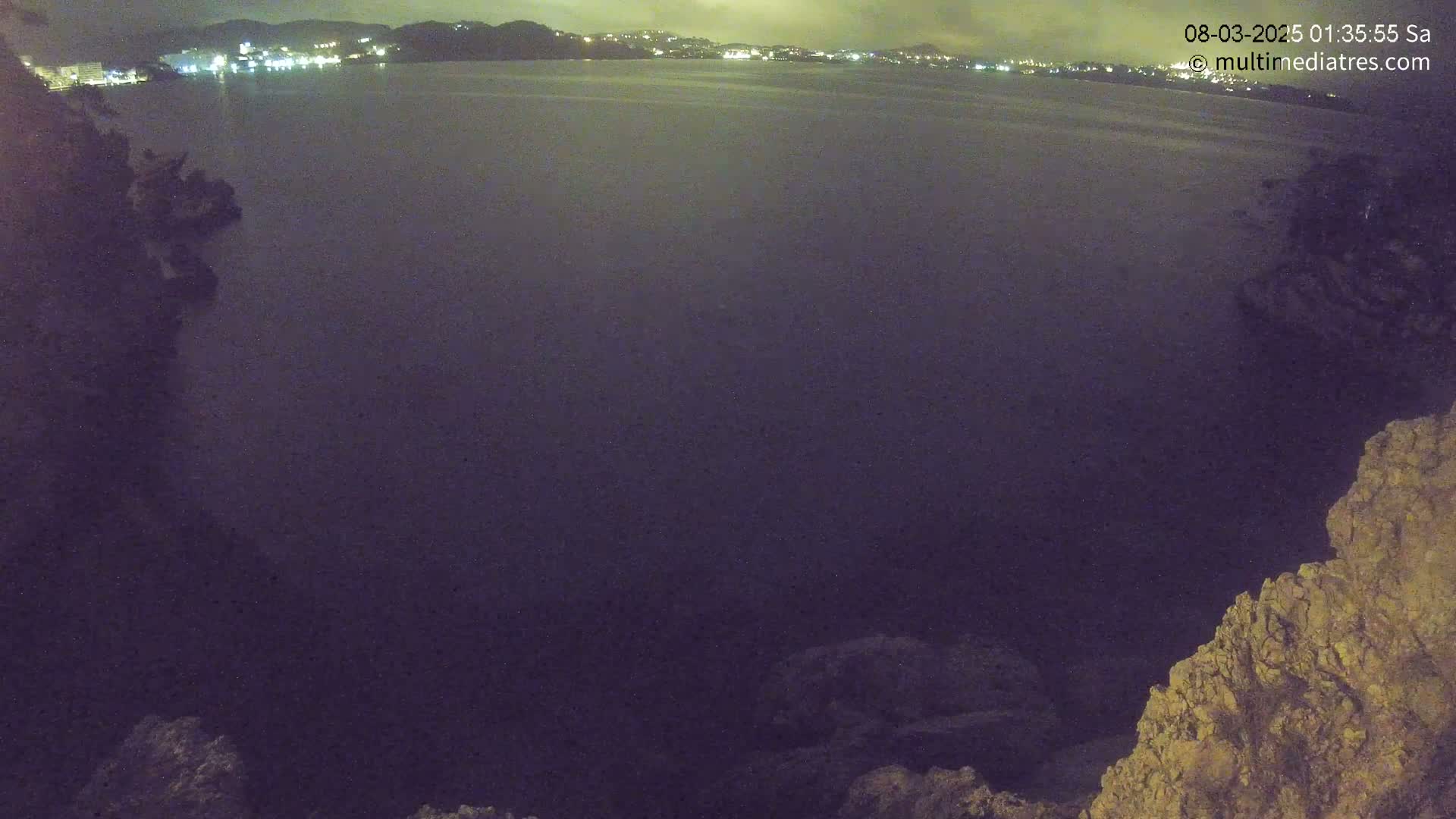 A nighttime, possibly overcast, view of a dark body of water with a distant town's lights visible across the water and rocky terrain in the foreground.