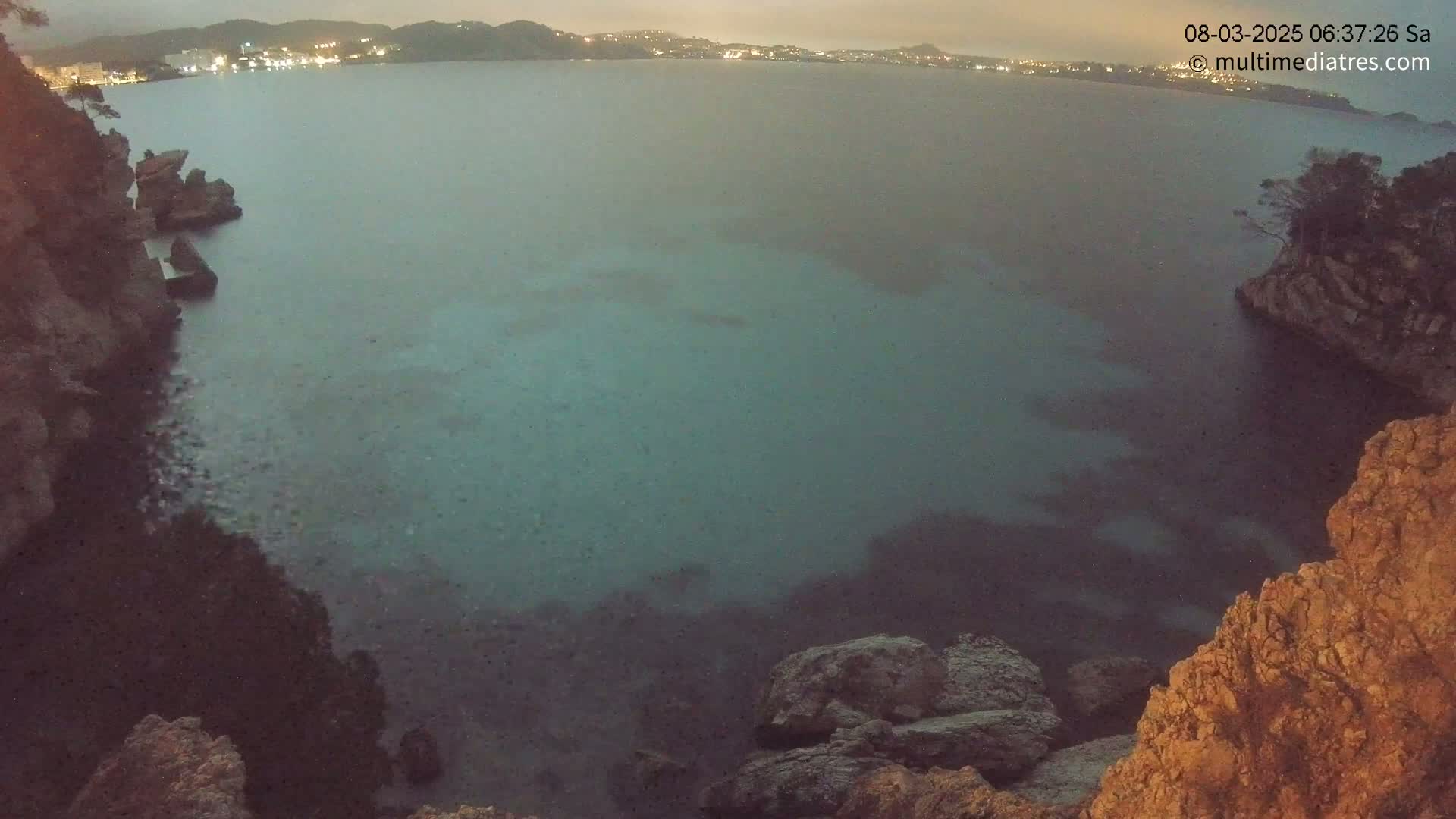 A dark, calm body of water is seen at dusk or night, nestled between rocky, cliff-like shores with a town's lights visible in the distance.
