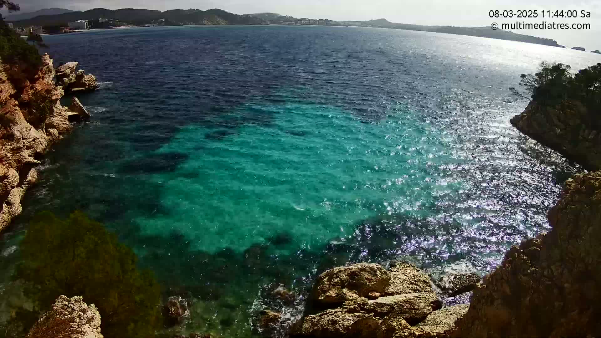 Sparkling turquoise water laps against rocky, tan-colored cliffs on a sunny day.