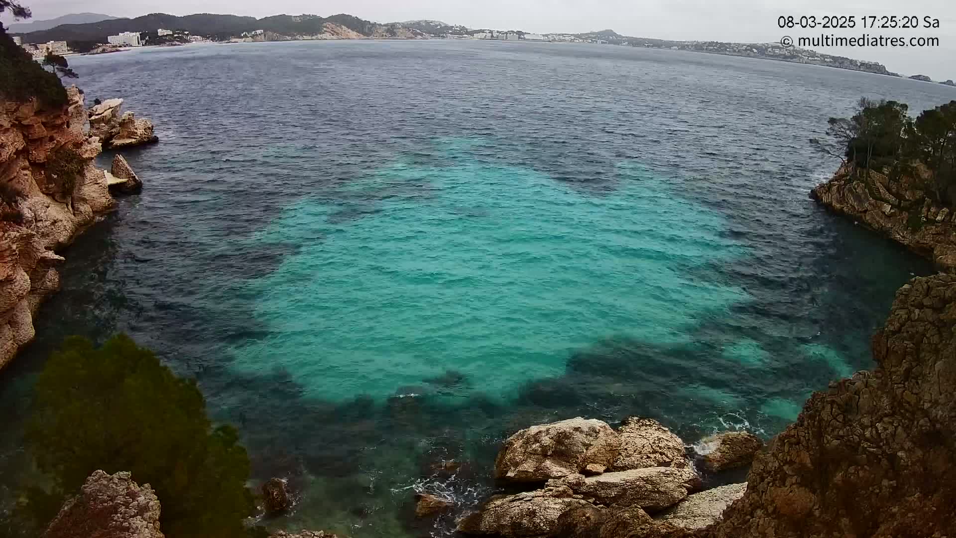 A cove of teal water is nestled between rocky cliffs, with a town visible in the distance under an overcast sky.