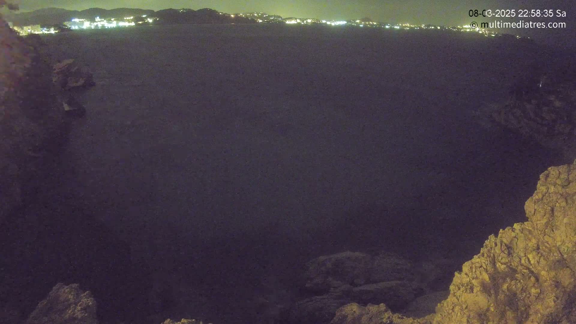 A nighttime view of a dark body of water with a distant city skyline illuminated, seen from a rocky outcrop.