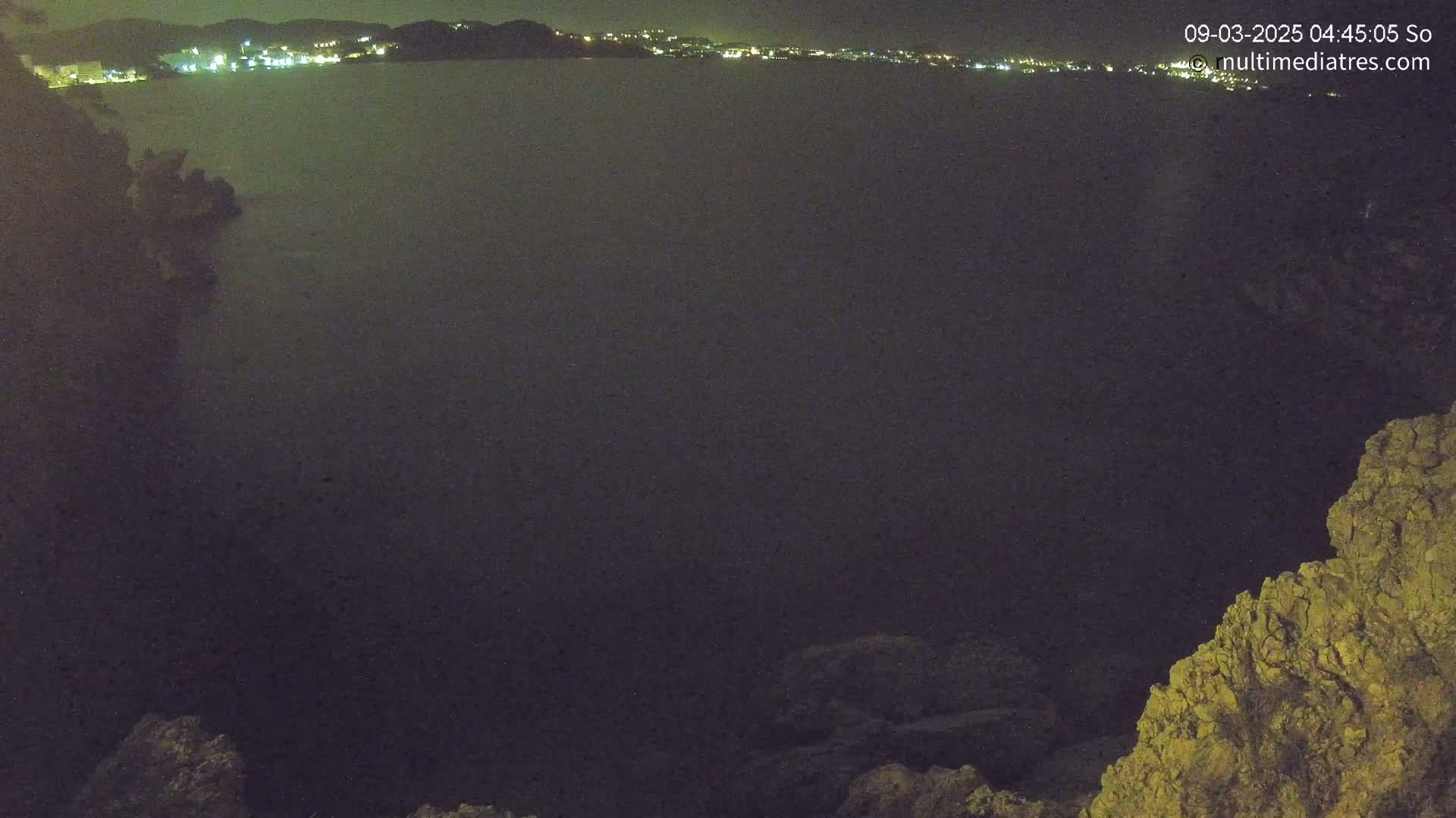 A nighttime view of a dark body of water with faintly lit buildings on the far shore and rocky terrain in the foreground.