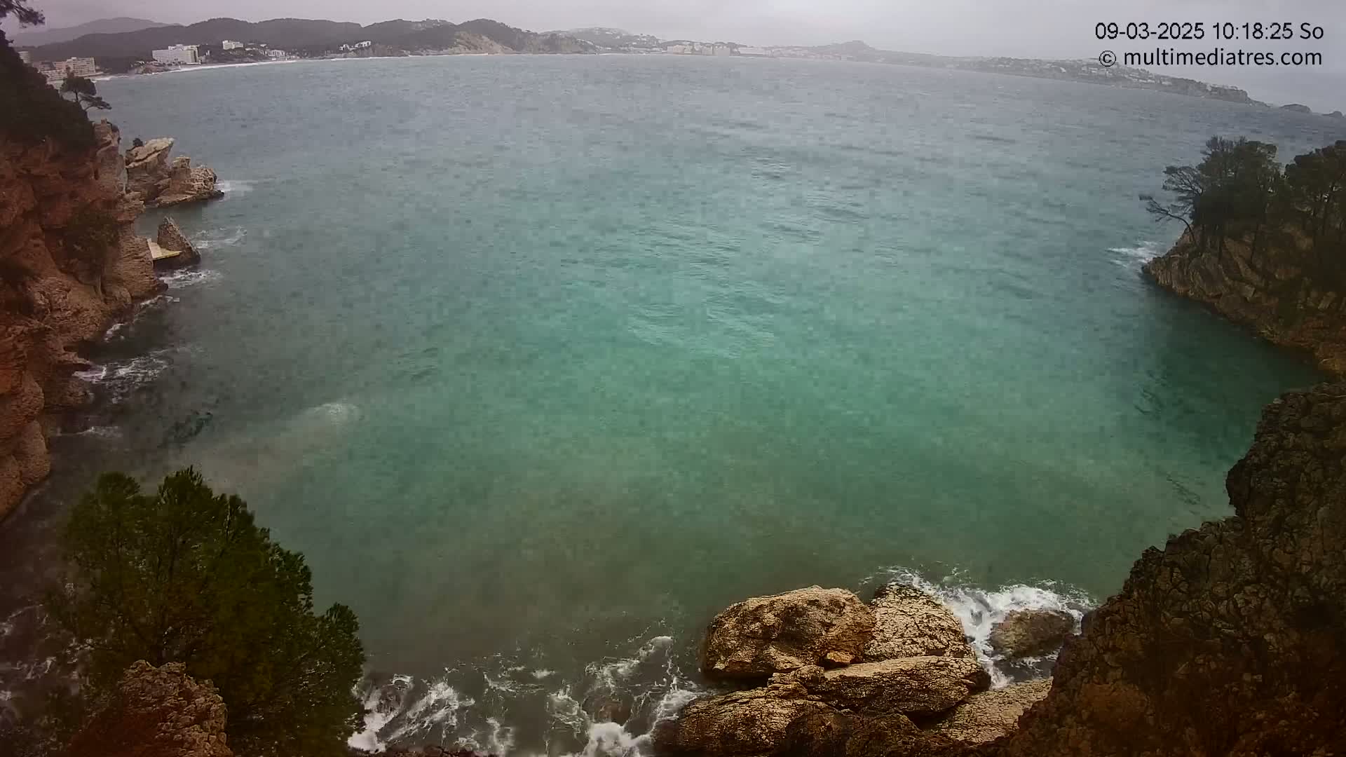 A teal-colored bay is seen from above, partially enclosed by rocky cliffs and a distant shoreline under an overcast sky.