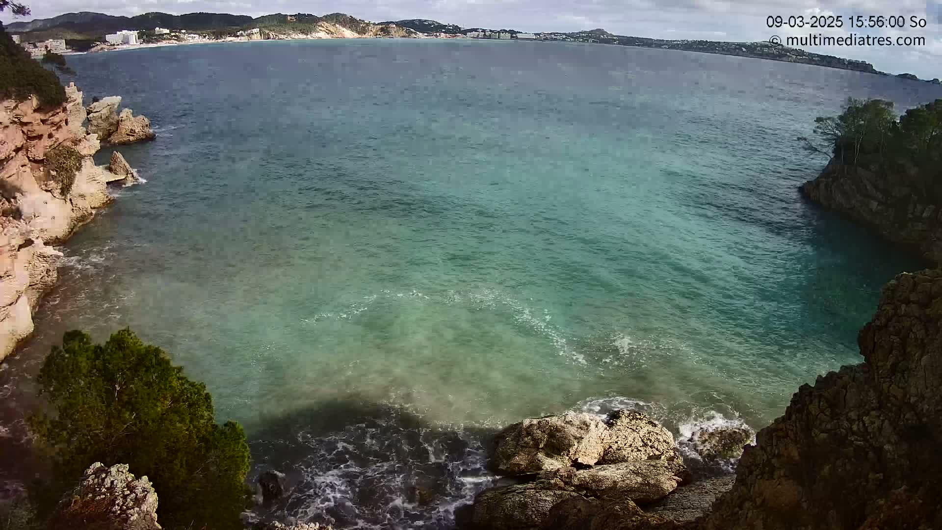 A cove of teal water is nestled between rocky cliffs, with a calm sea and distant coastline visible under a partly cloudy sky.