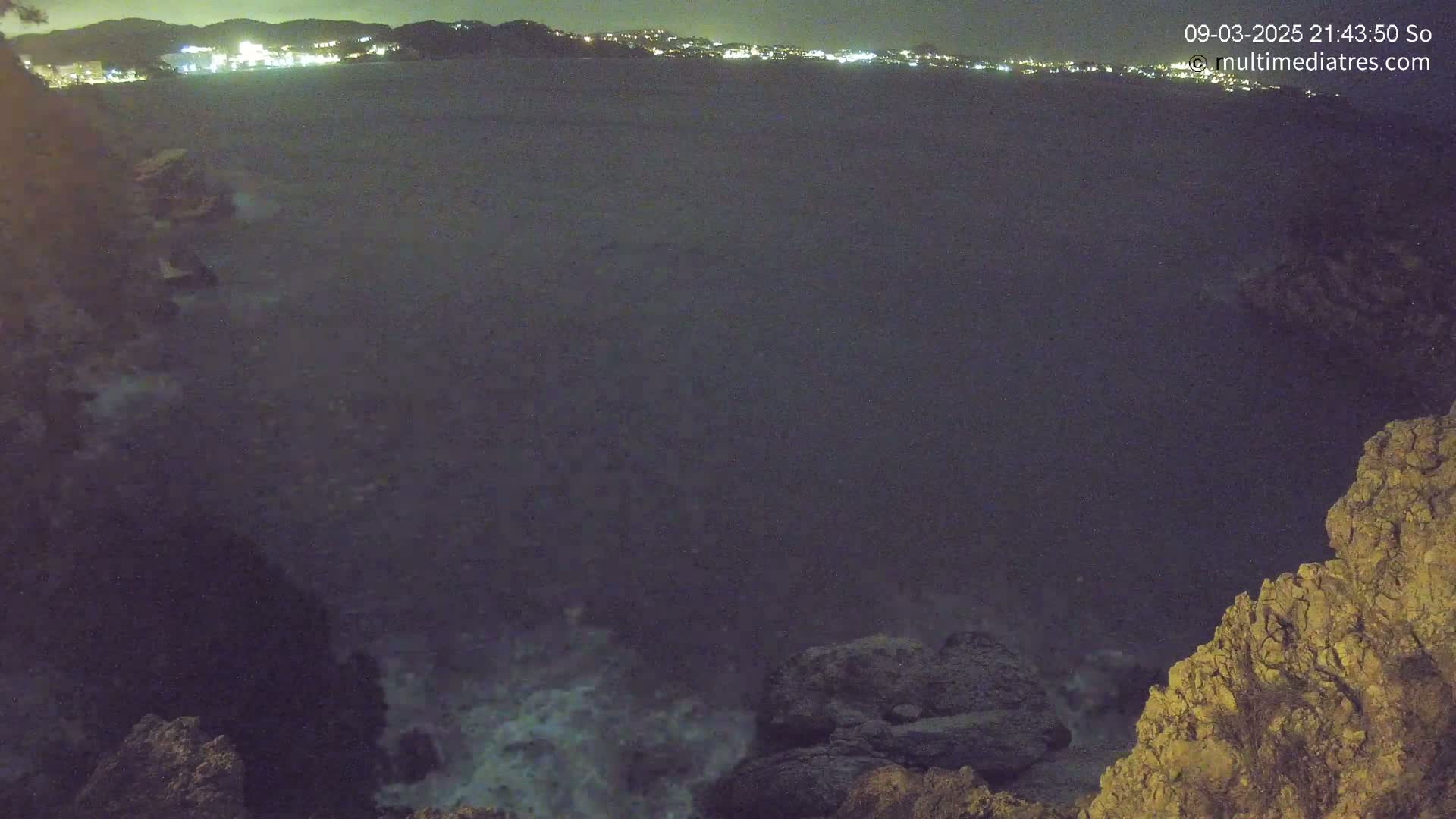 A nighttime view of a dark body of water with waves crashing against rocky terrain, a city's lights visible in the distance.