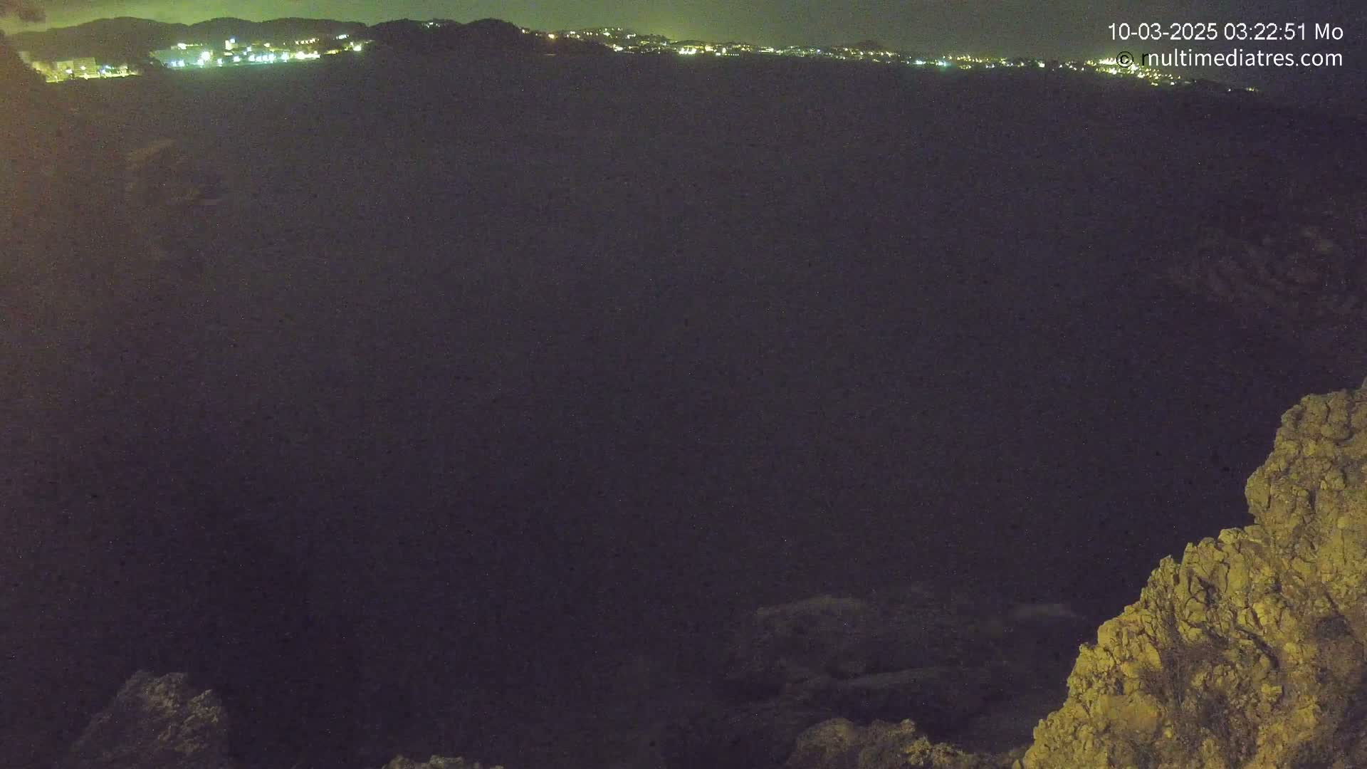 A nighttime view of a dark, possibly volcanic, landscape with a distant line of city lights.