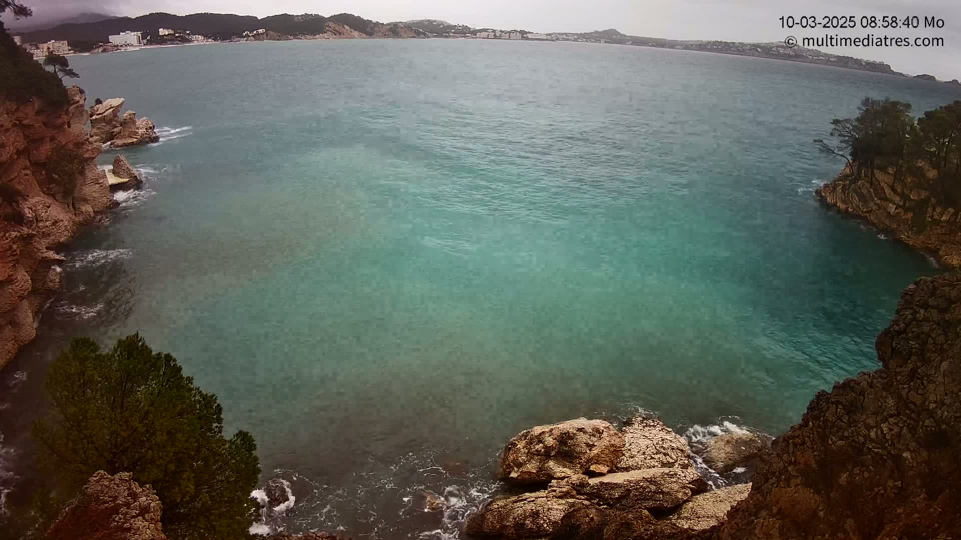 A rocky cove with teal water meets a calm sea under an overcast sky, with a distant coastline visible in the background.