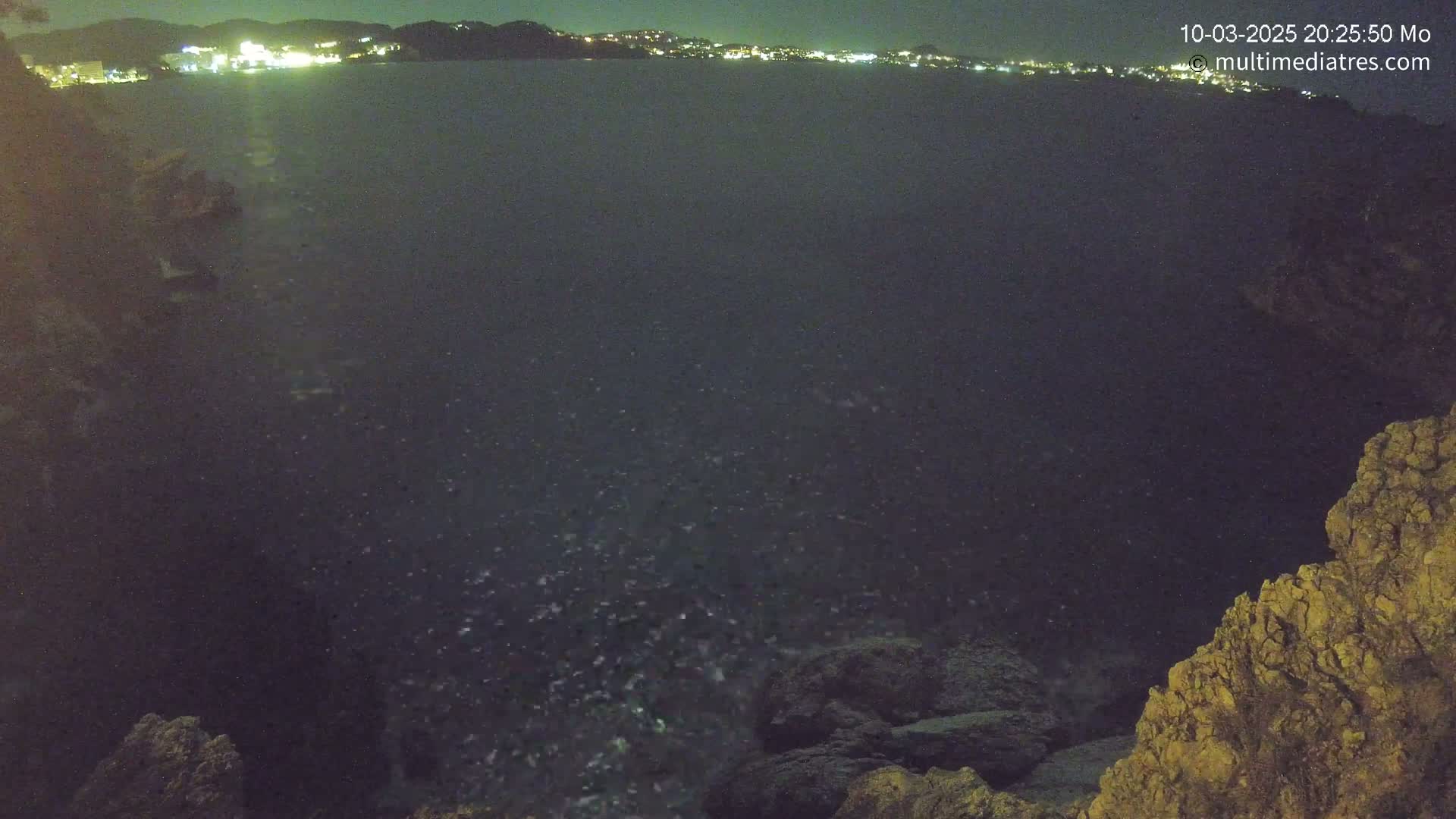 A dark, nighttime view of a rocky shoreline overlooking a body of water with a distant city illuminated at night.