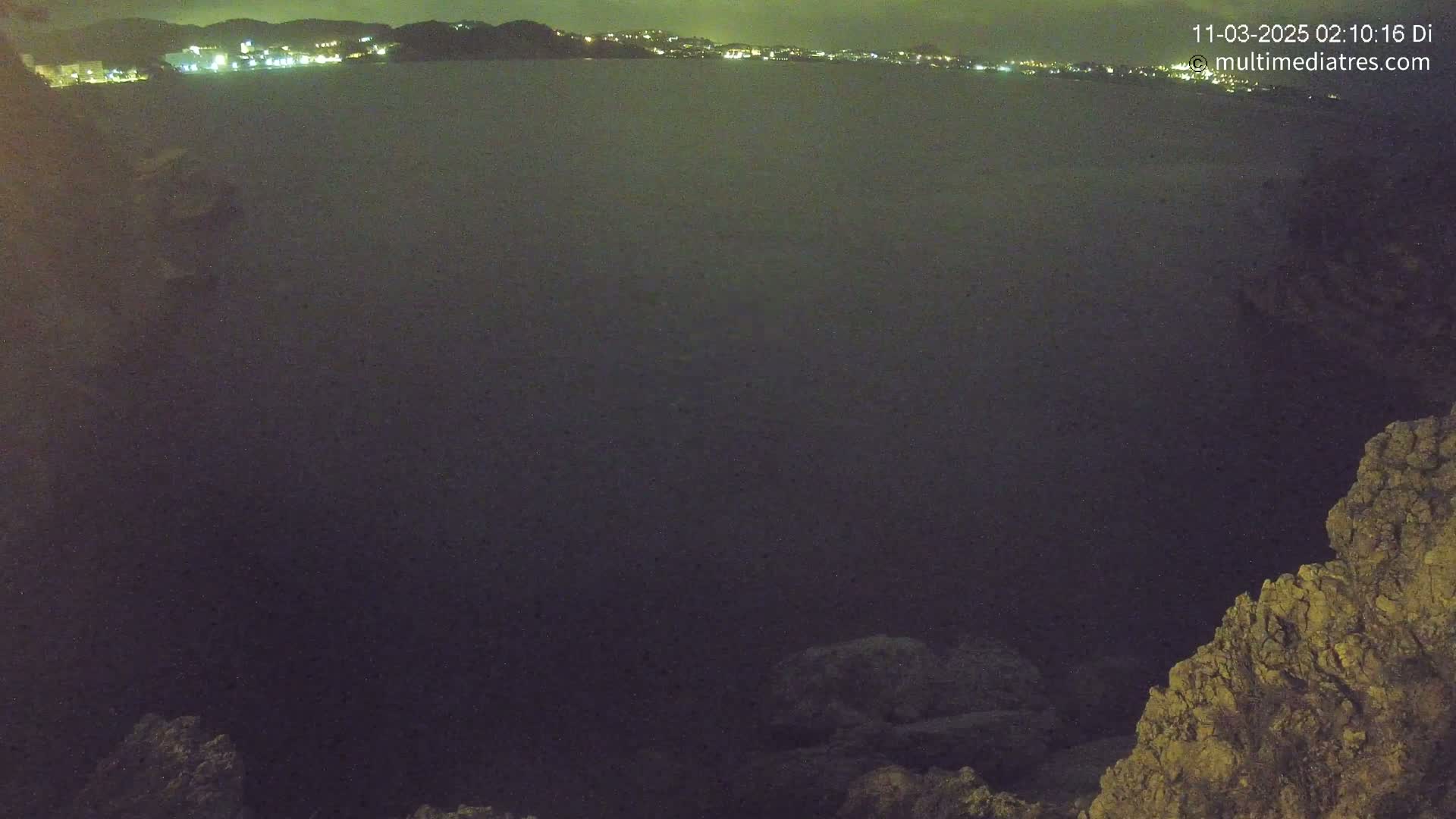 A nighttime view of a dark body of water with a distant, faintly lit coastline under a cloudy sky.
